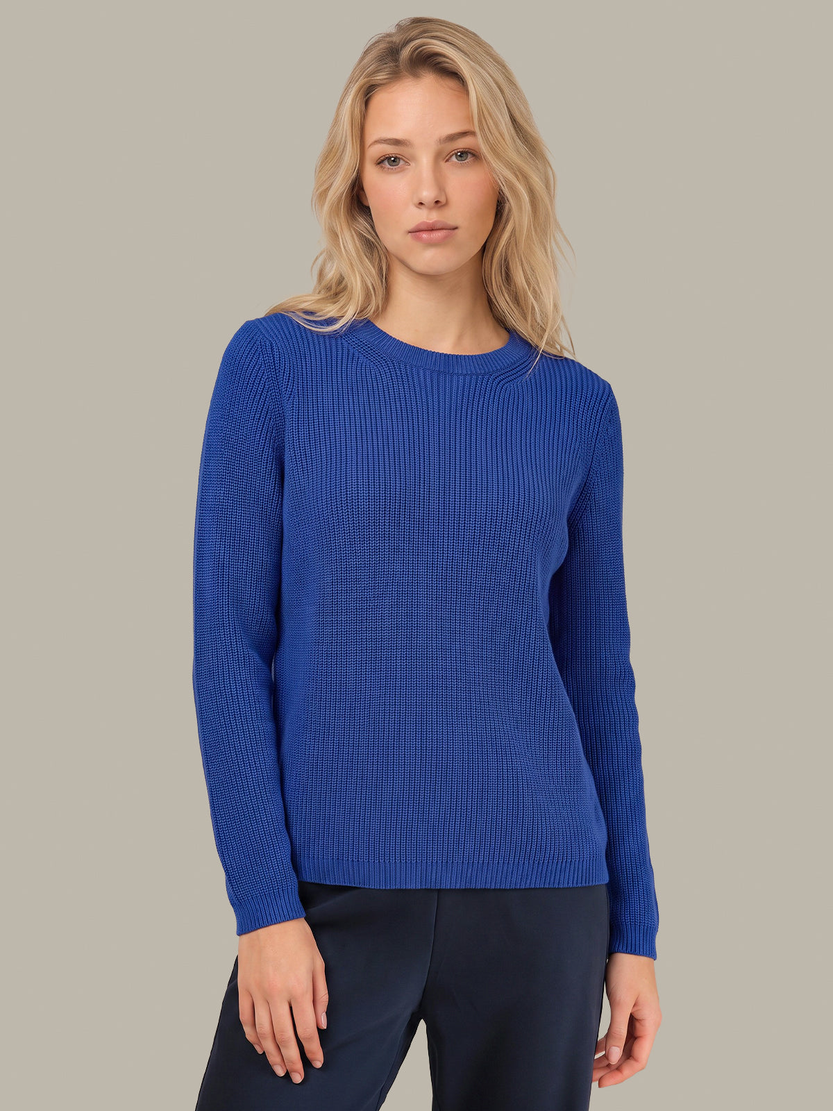 A blonde woman wearing a royal blue ribbed knit long-sleeve sweater and dark navy pants, standing against a solid light gray studio background.
