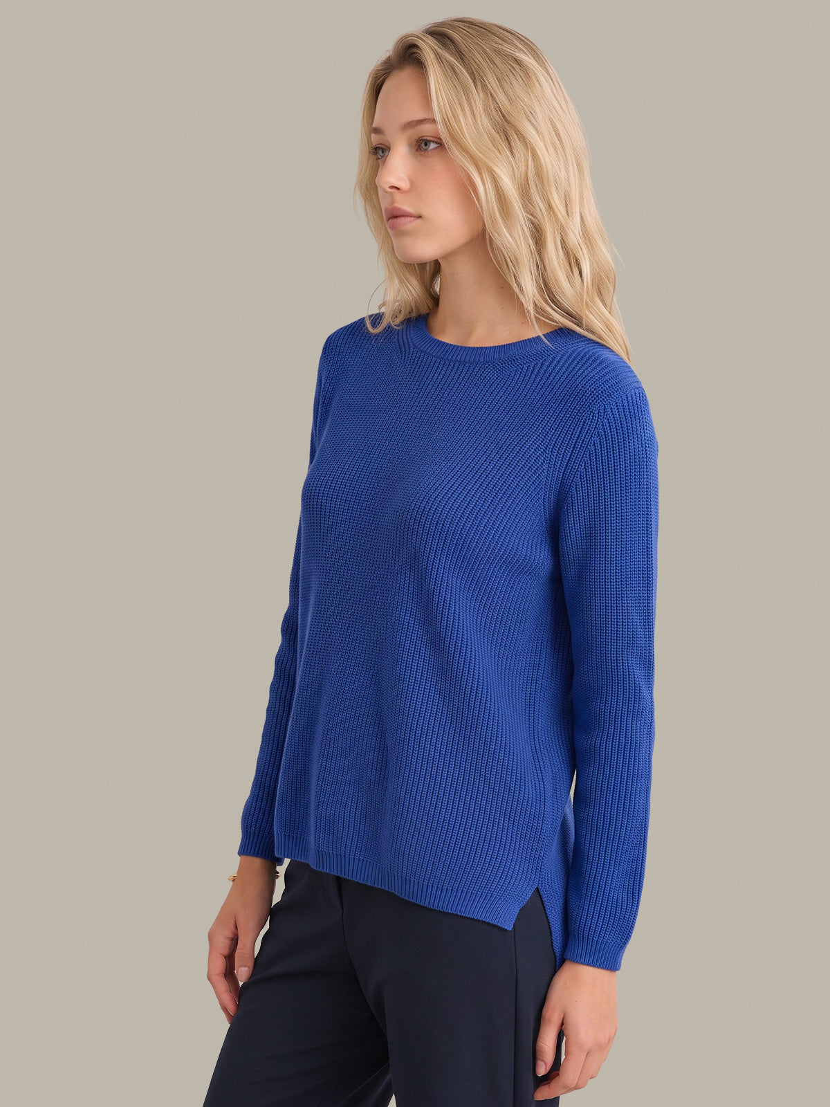 A woman wearing a royal blue ribbed knit sweater and dark trousers, photographed against a solid light gray studio background.