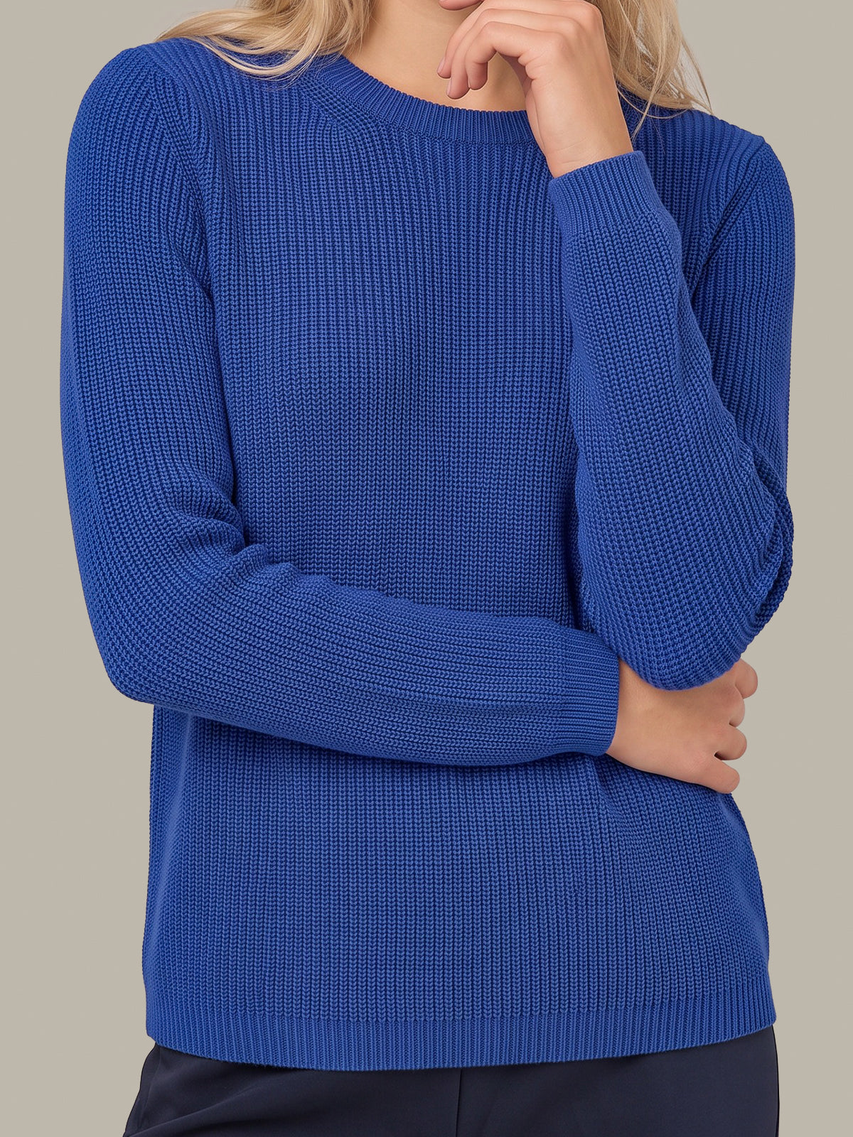A woman wearing a royal blue ribbed knit sweater with long sleeves and a crew neckline, shown from the shoulders to the hips against a plain warm gray background.