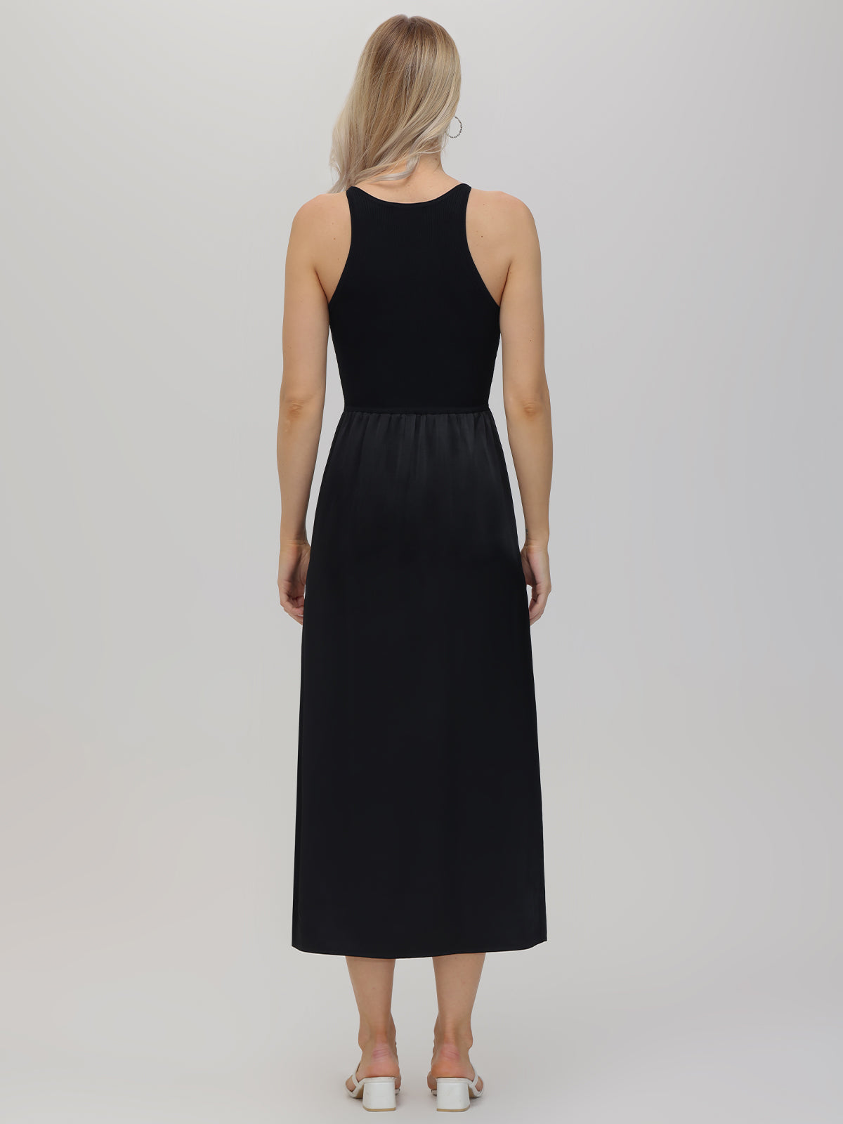 A person with long hair stands against a plain background, donned in a sleeveless, ankle-length black midi dress by 525 America and white sandals, ideal for semi-formal events.
