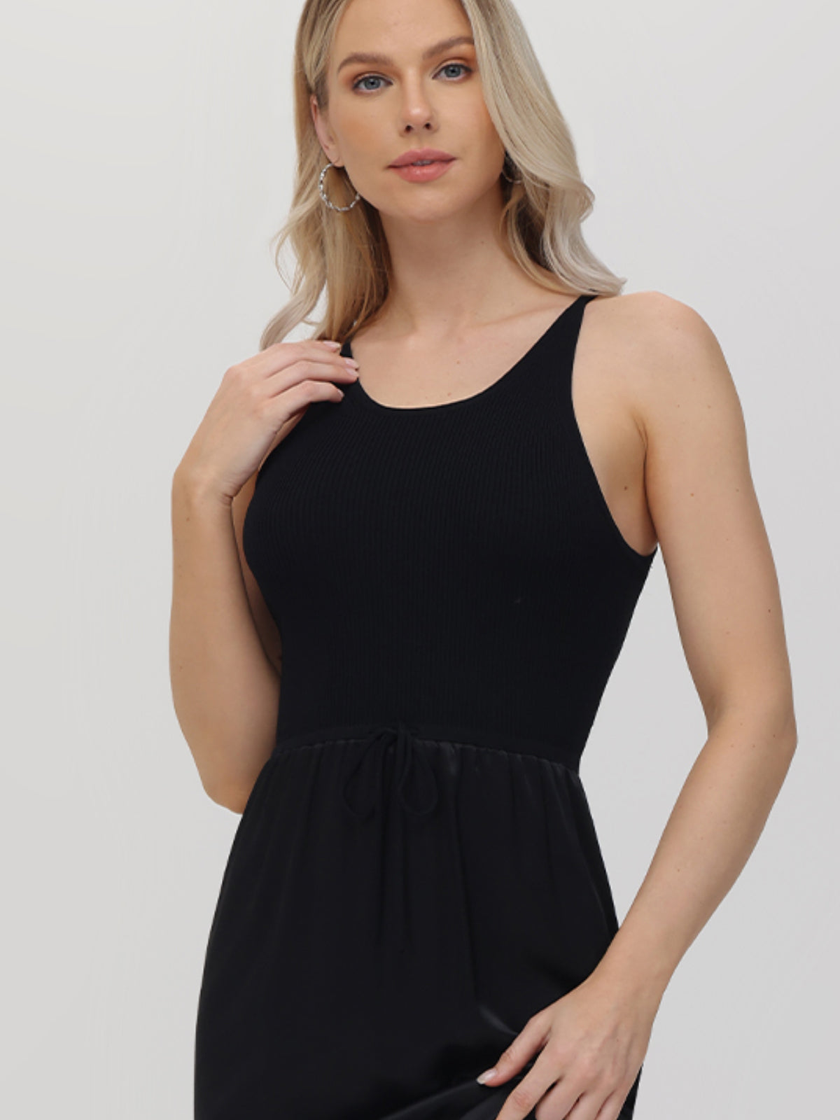 A long-haired blonde woman is wearing a sleek black sleeveless midi dress by 525 America, featuring a round neckline and fitted top, ideal for semi-formal events. She touches her hair with one hand while looking at the camera against a light-colored plain background.