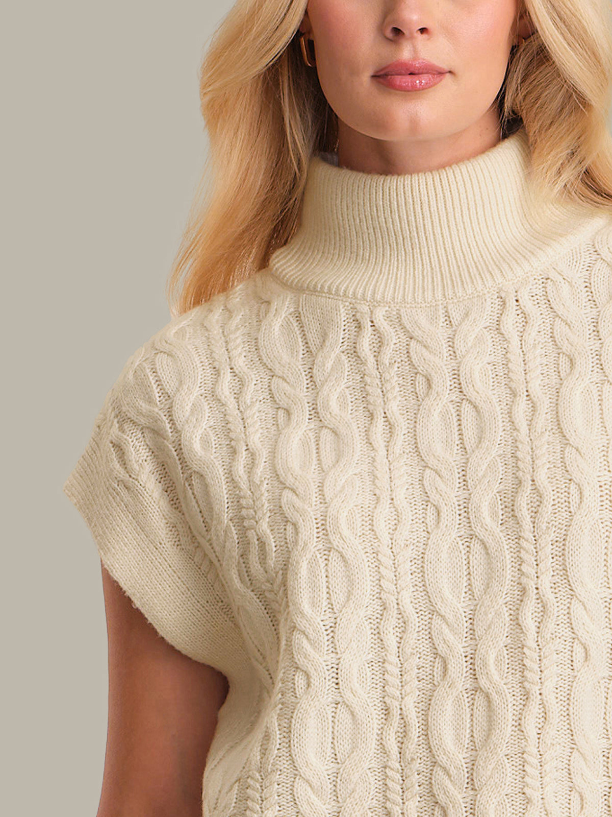 Close-up of a person wearing a beige cable knit sweater against a neutral background