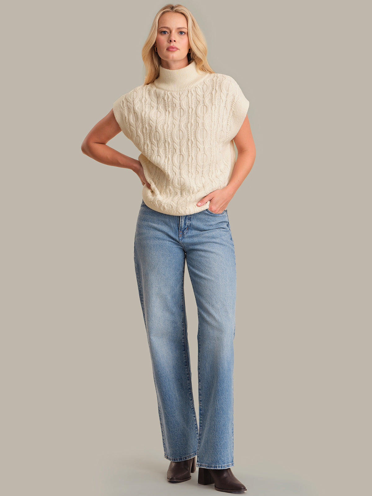 Woman wearing a cream sweater and blue jeans against a gray background