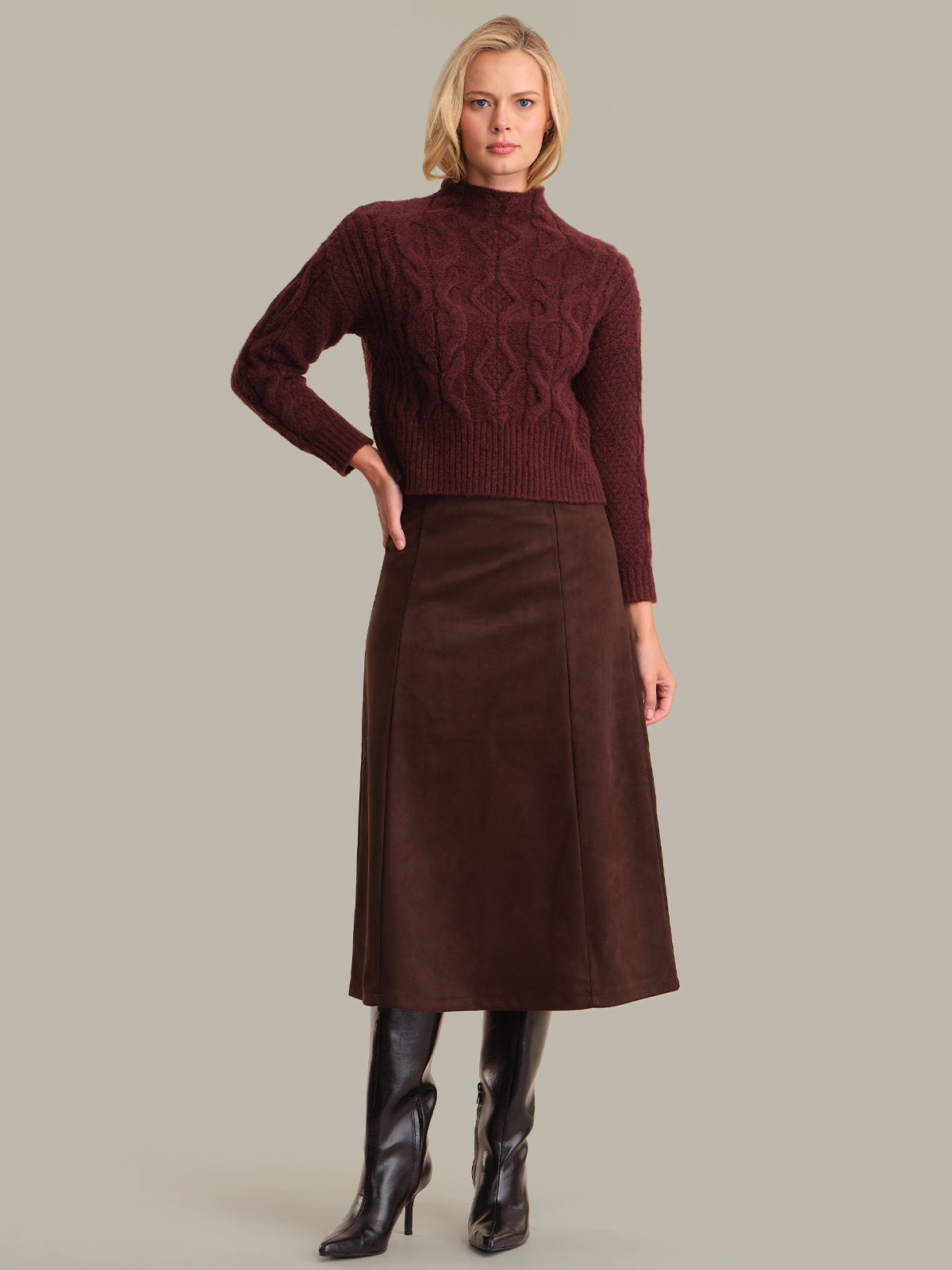 Woman wearing a burgundy cable knit sweater and brown skirt on a gray background