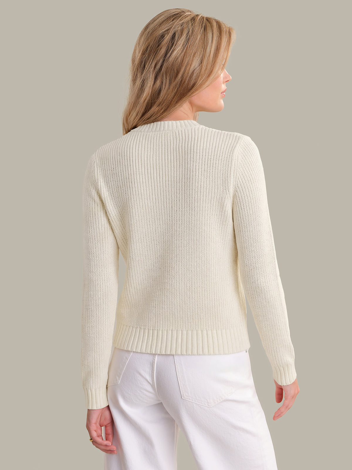 Woman wearing a cream sweater and white pants against a plain background