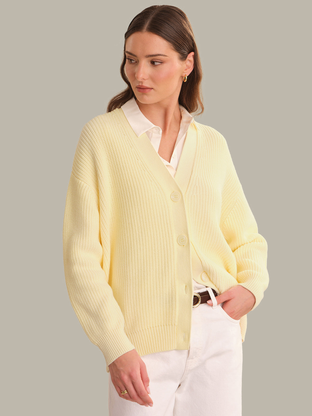 A woman wearing a pale yellow ribbed knit cardigan over a white collared shirt and white pants, standing against a solid light gray background.