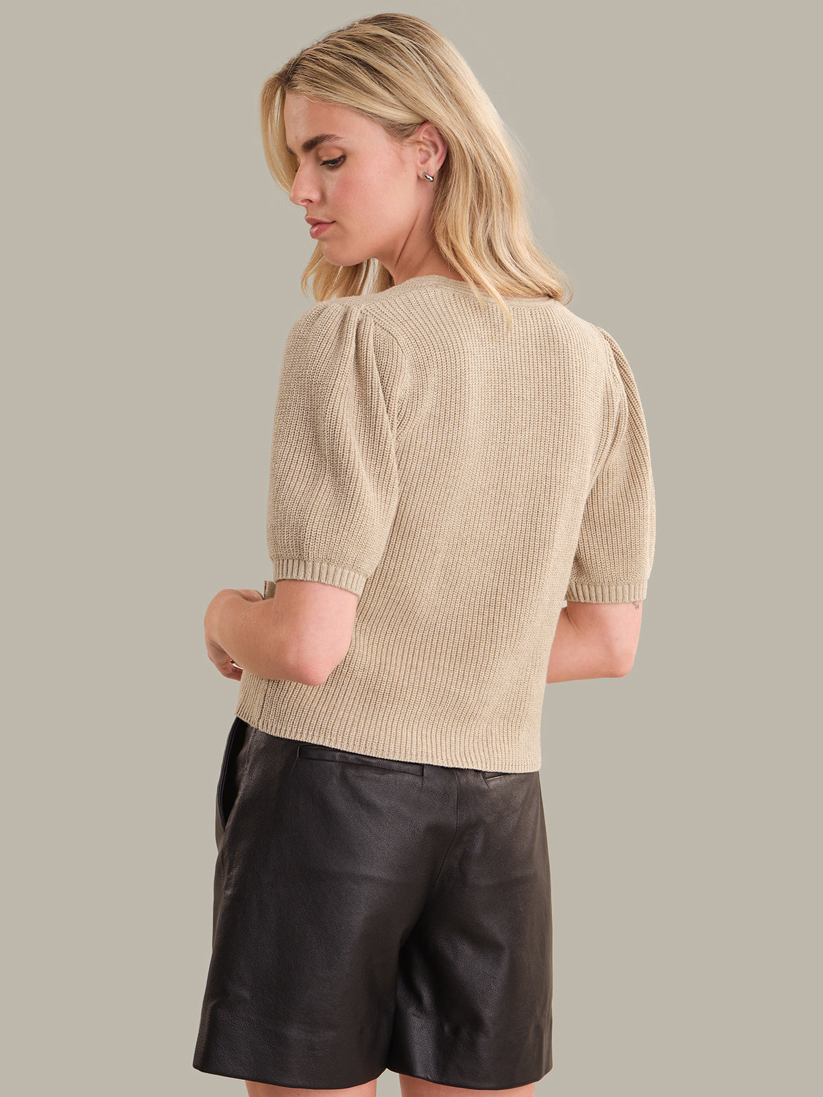 Woman wearing a beige knit top and black leather shorts on a plain background