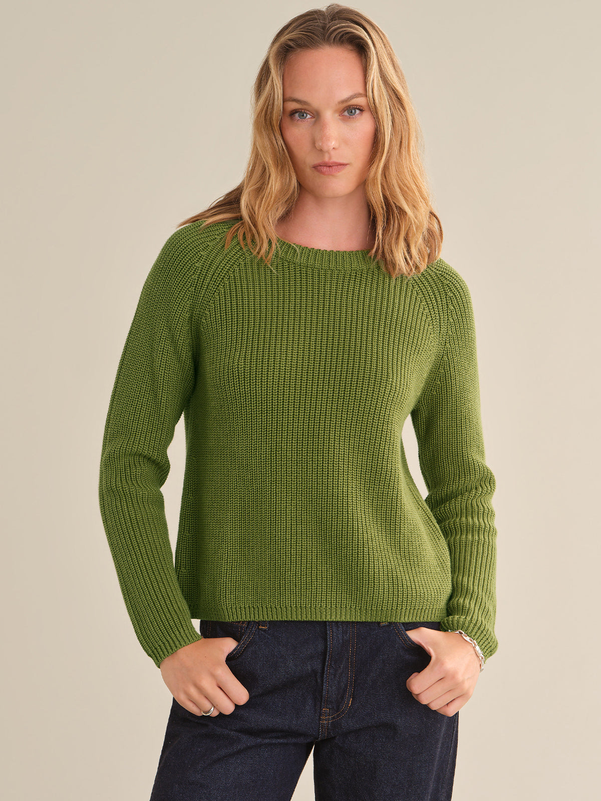 A woman wearing a ribbed knit crewneck sweater in olive green, paired with dark blue jeans, standing against a solid light beige background.