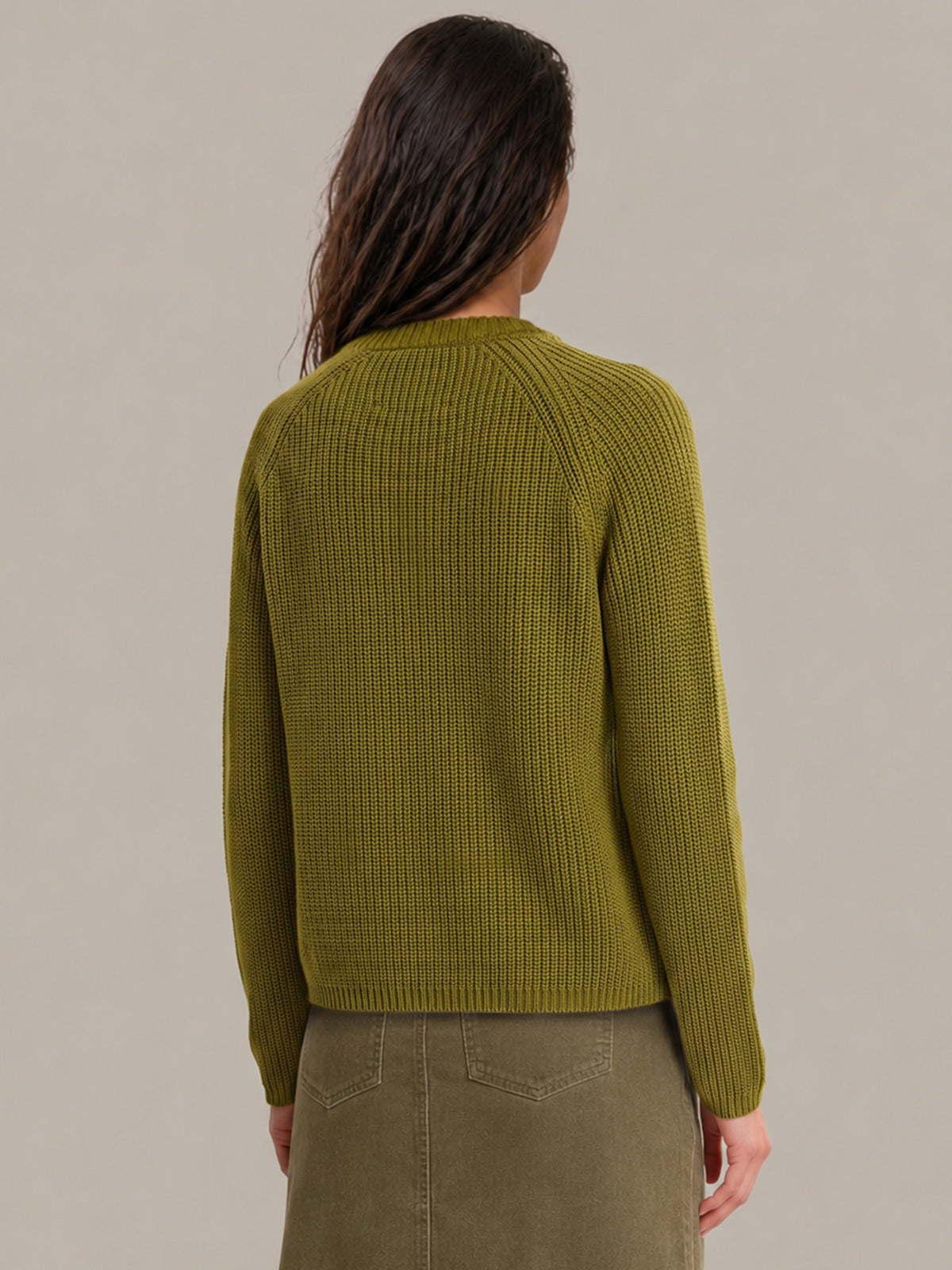 A woman viewed from the back wearing a ribbed knit long-sleeve sweater in olive green, paired with an olive green skirt, against a solid light gray background.