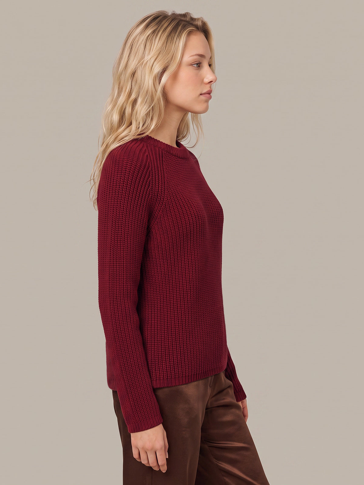 A woman in profile wearing a ribbed long-sleeve crewneck sweater in deep burgundy, paired with brown pants, against a solid light gray studio background.