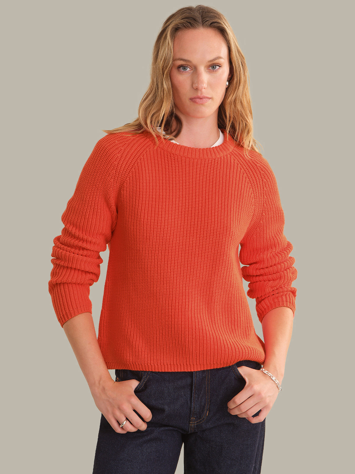 A woman wearing a ribbed knit crewneck sweater in a vibrant orange color, paired with dark blue jeans, standing against a solid light gray background.
