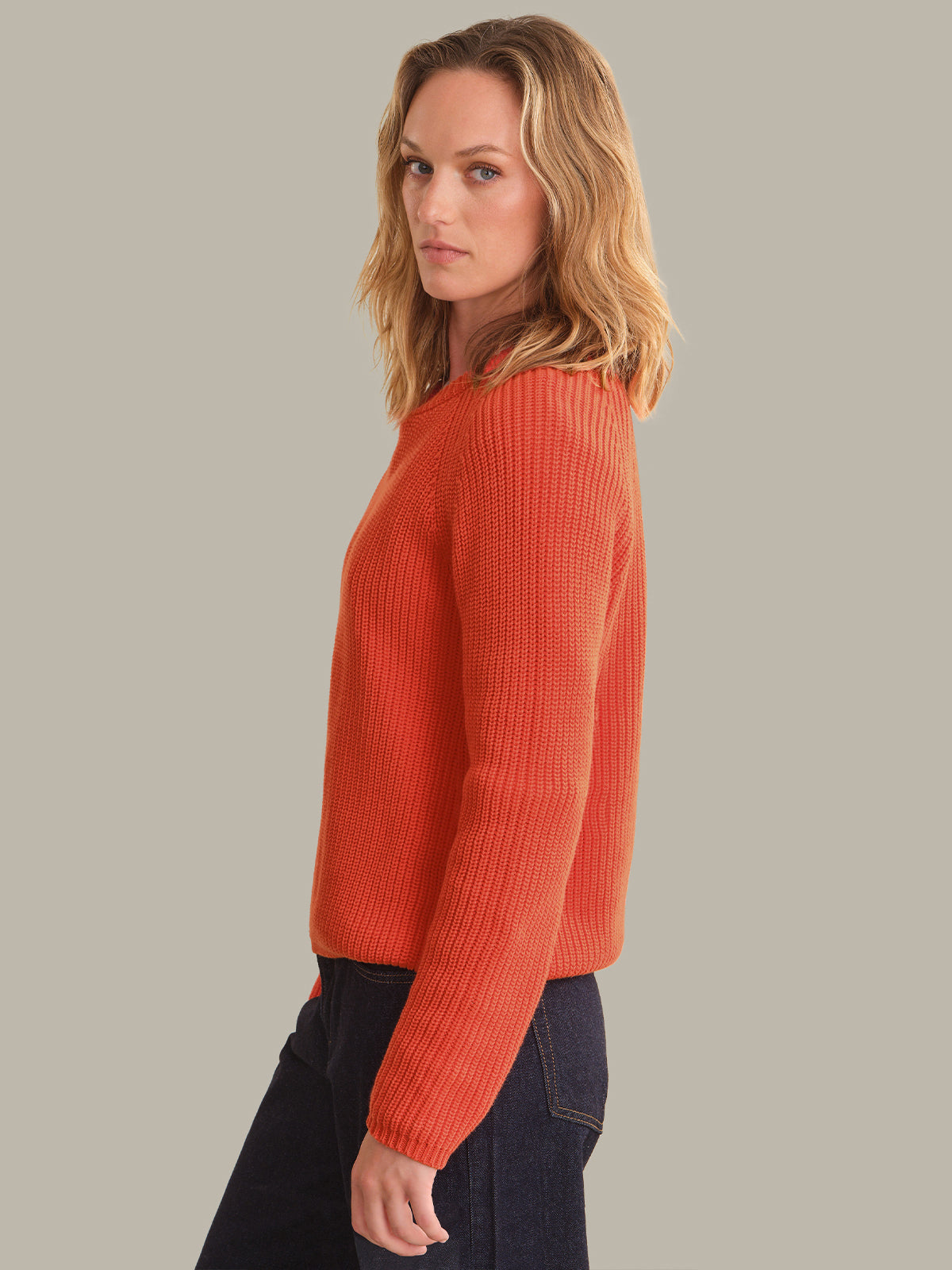 A woman wearing a bright orange ribbed knit sweater and dark blue jeans, shown from a side profile against a solid light gray background.