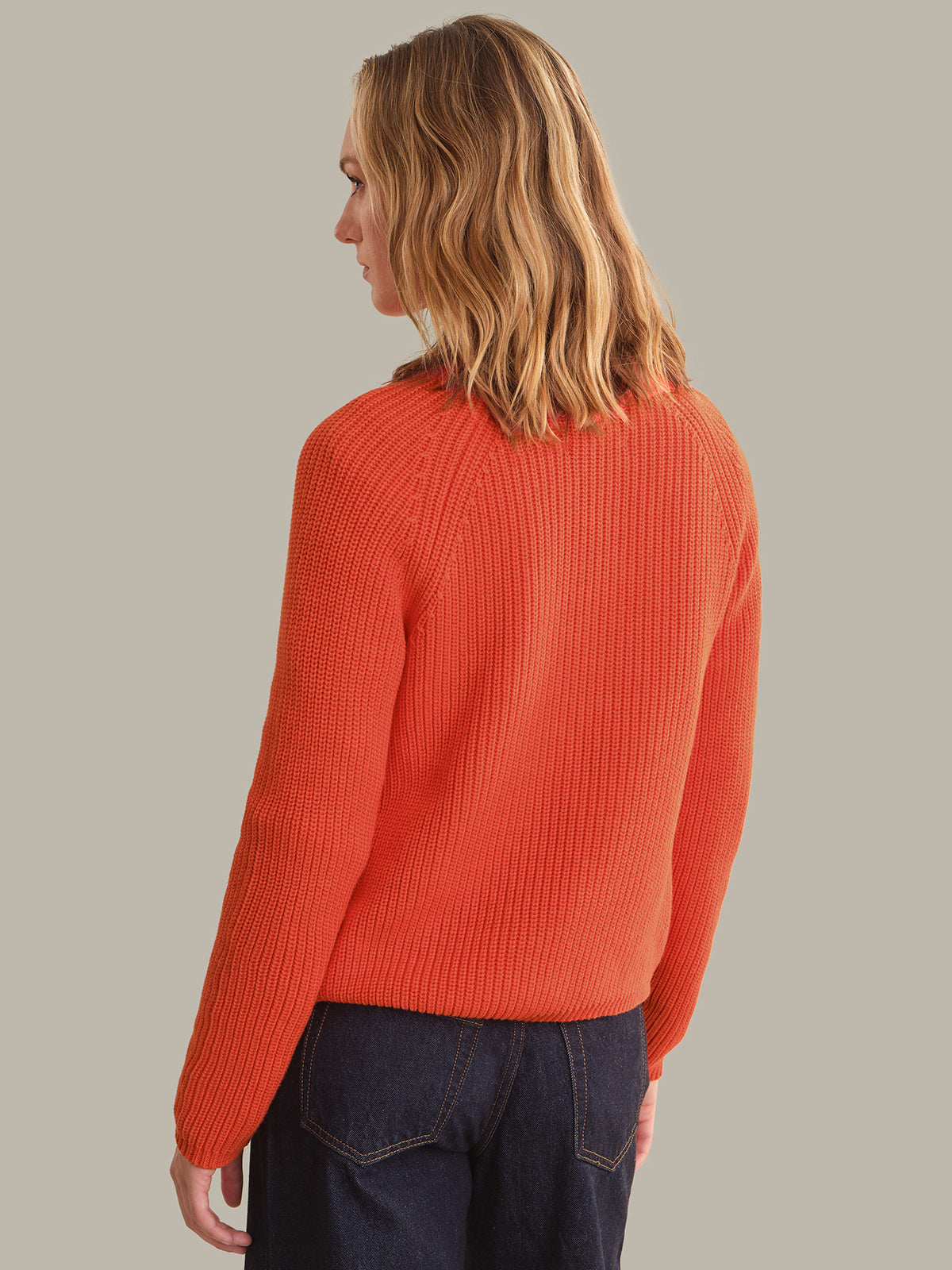 A woman with blonde hair is shown from the back wearing a bright orange ribbed knit sweater and dark blue jeans, set against a solid light gray background.