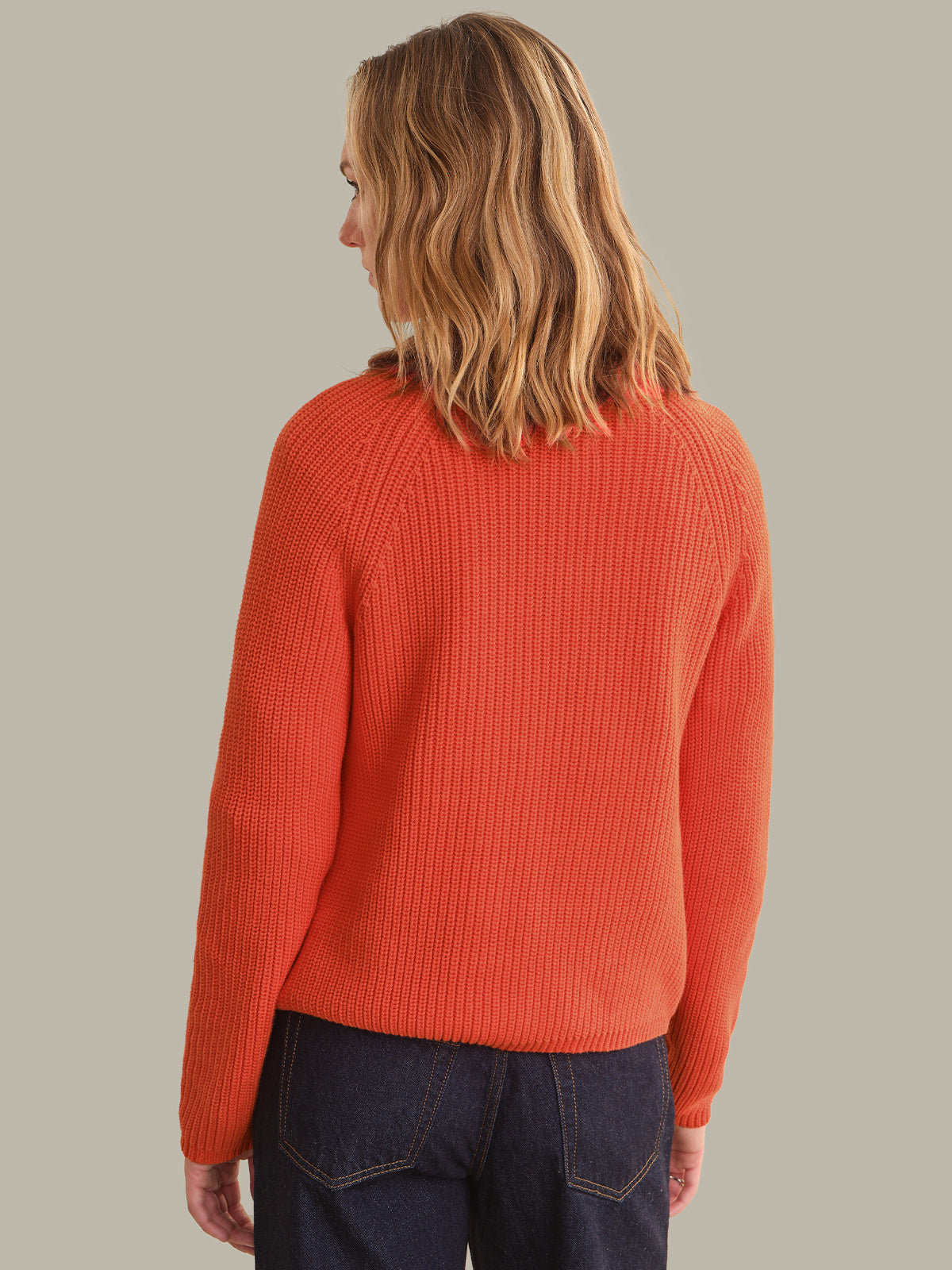 A woman viewed from the back wearing a bright orange ribbed knit sweater and dark blue jeans, set against a solid light gray background.
