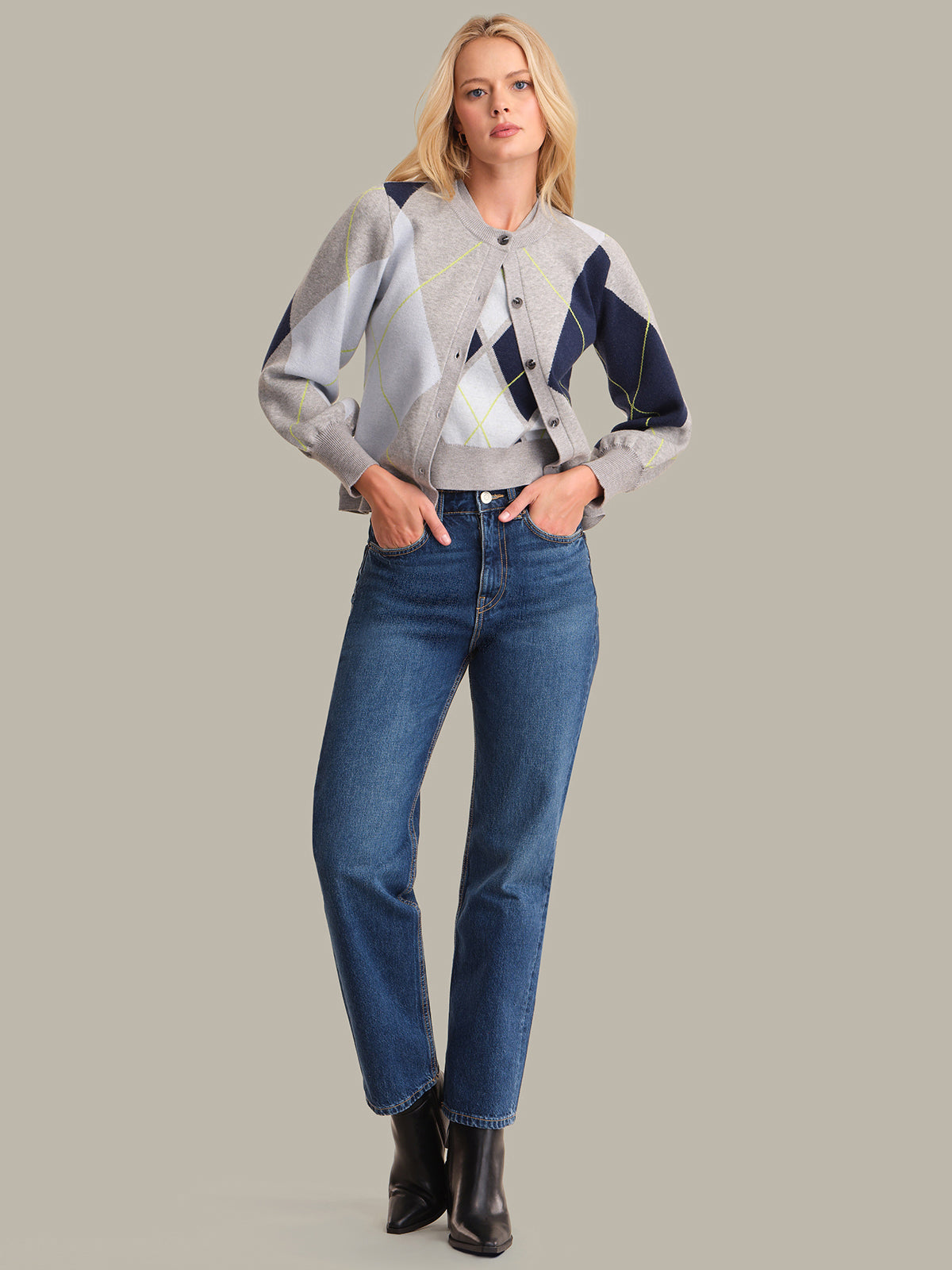 Woman wearing a patterned jacket, blue jeans, and black boots on a gray background