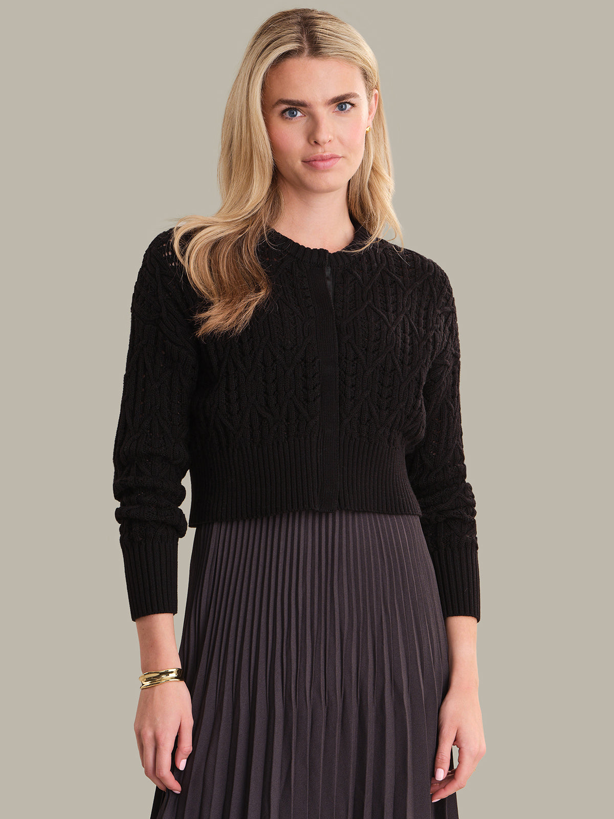 Woman wearing a black cable knit sweater and dark pleated skirt on a gray background