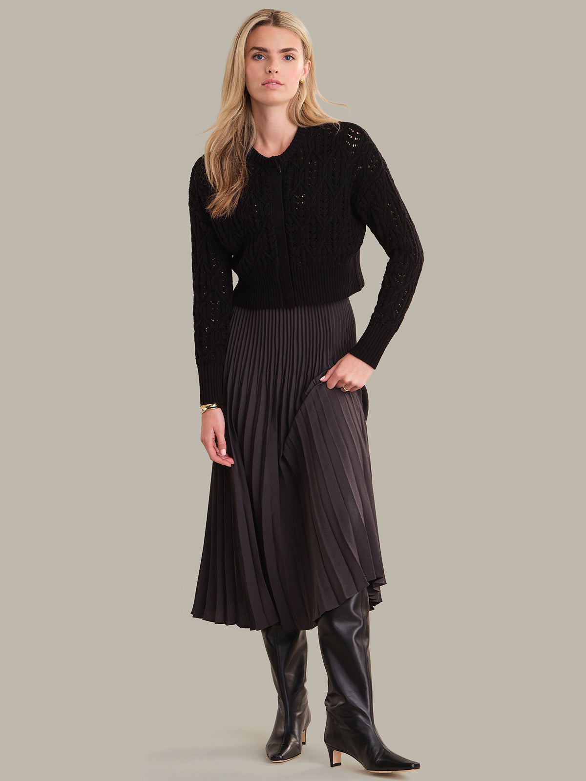 Woman wearing a black sweater and pleated skirt with black boots on a gray background