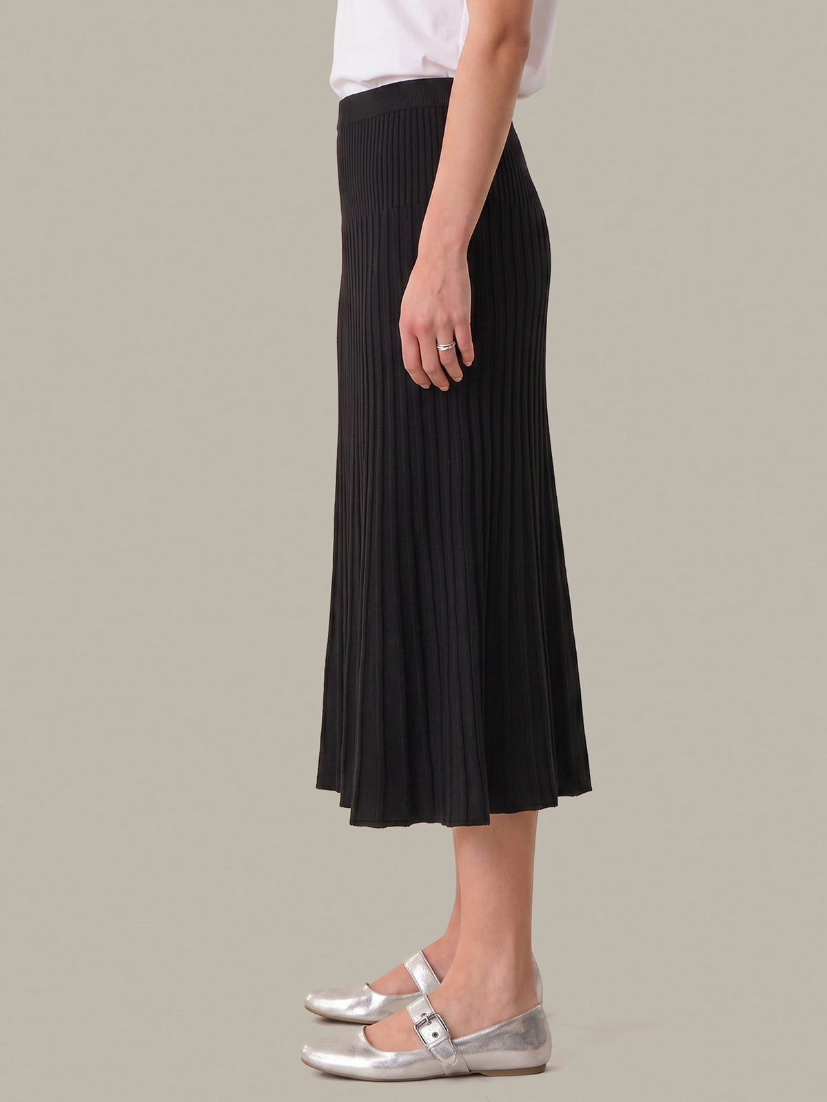 A side-profile view of a model wearing a black pleated midi skirt, paired with a white top and metallic silver Mary Jane flats, set against a solid light gray background.