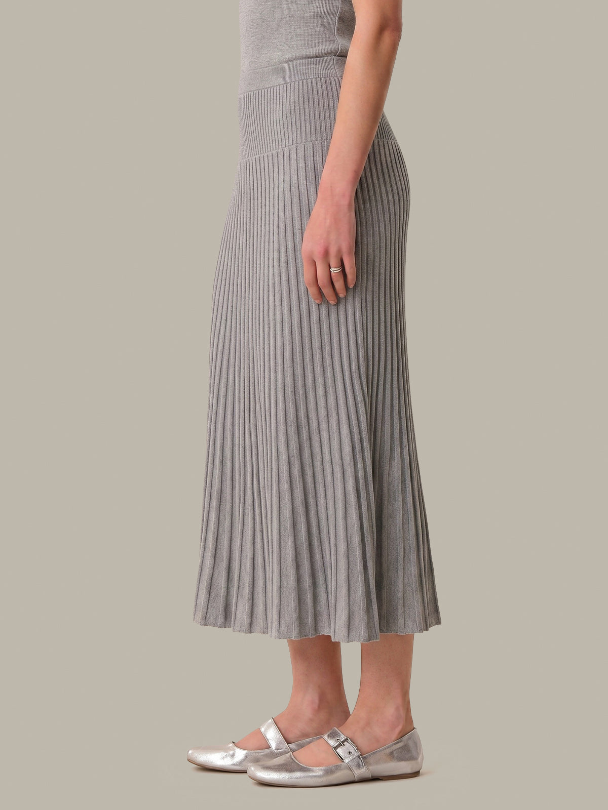 A model wearing a gray pleated midi skirt, a matching gray ribbed top, and metallic silver Mary Jane flats, shown from the waist down against a solid light gray background.