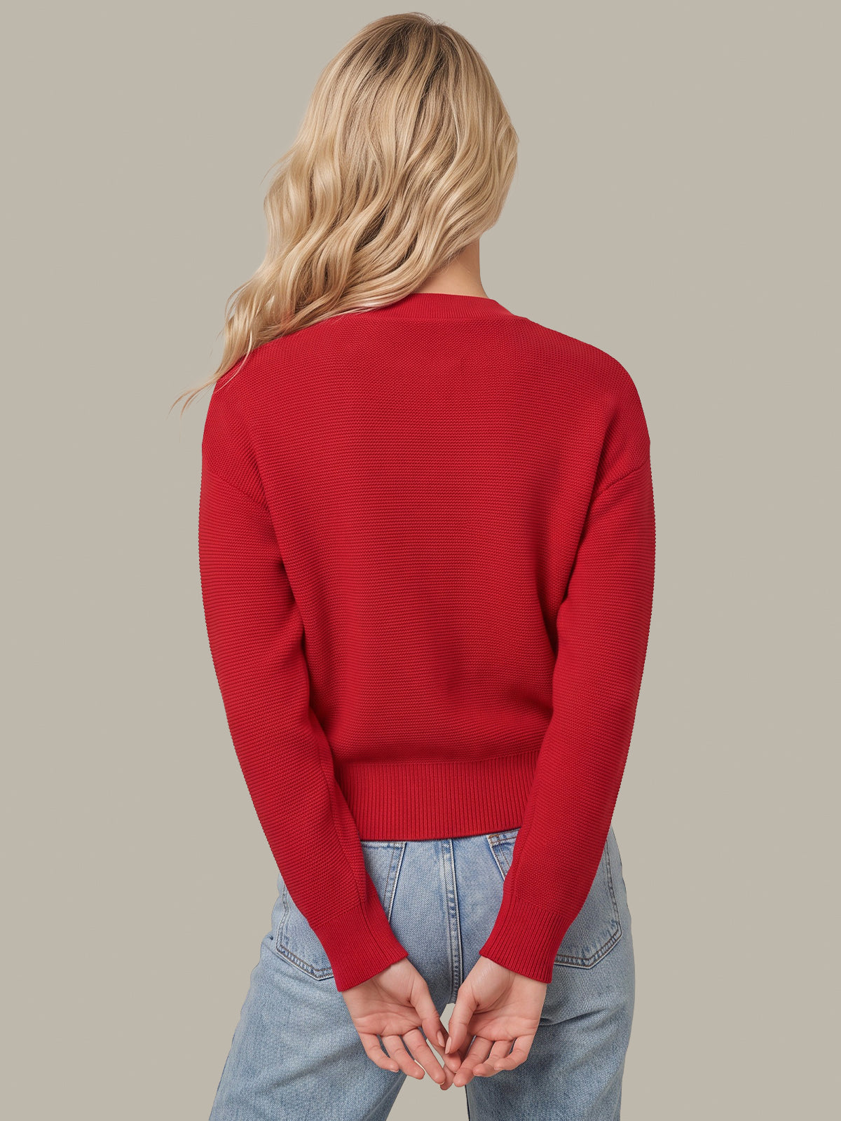 A woman with blonde hair is shown from behind wearing a bright red knit sweater and light blue jeans against a solid gray background.