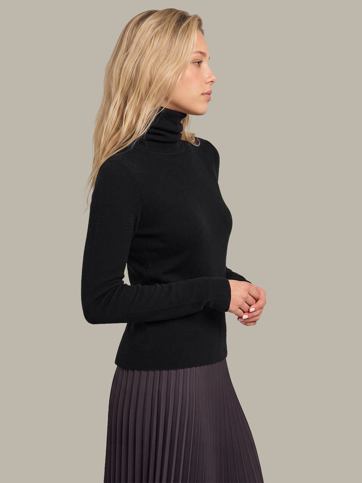A woman in profile wearing a black long-sleeve turtleneck sweater and a dark pleated skirt, photographed against a plain warm gray studio background.