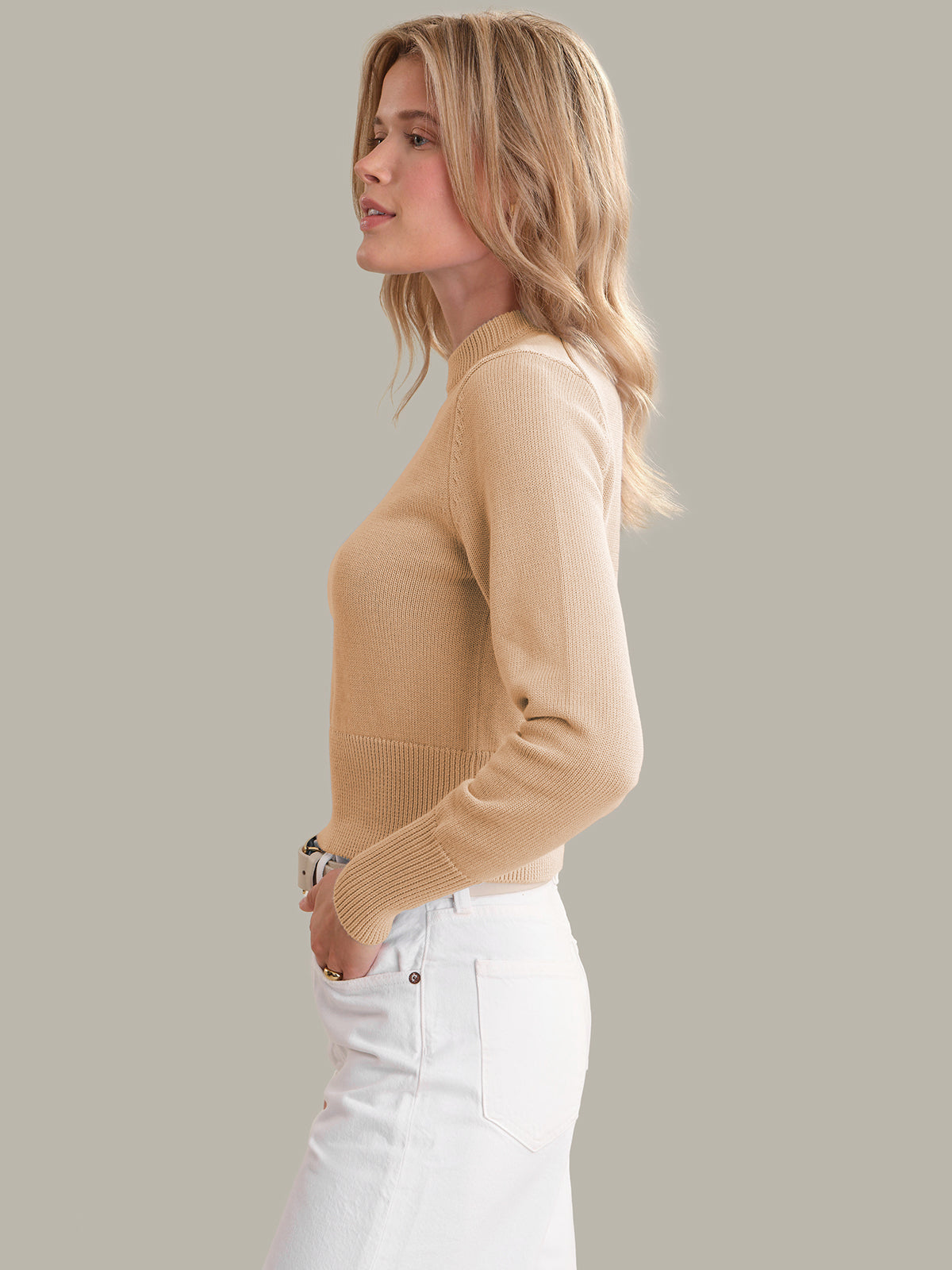 Woman wearing a beige sweater and white pants against a plain background