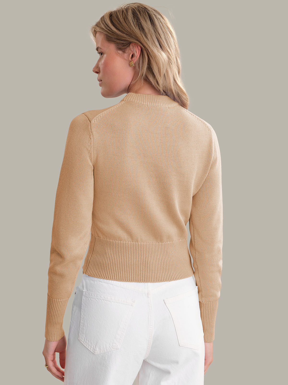 Woman wearing a beige sweater and white pants against a plain background