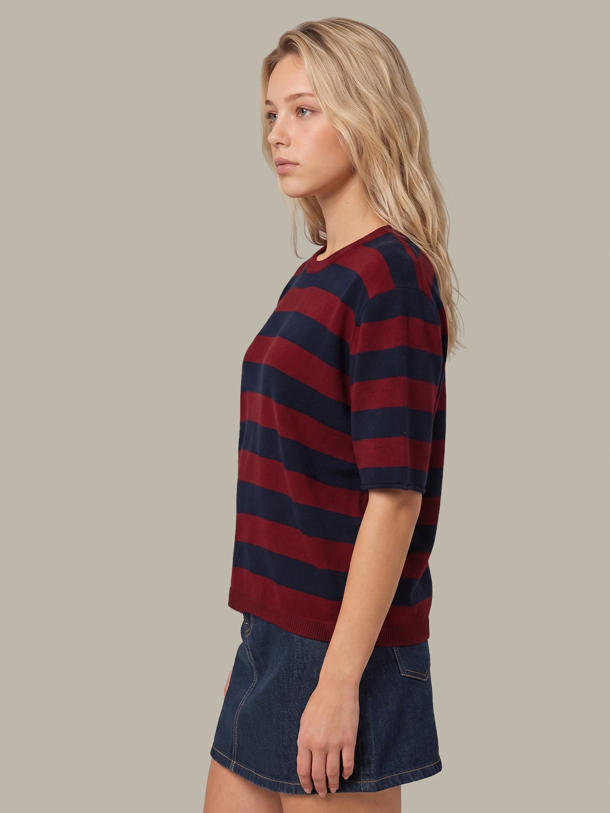 A woman shown in side profile wearing a short-sleeve crewneck t-shirt with horizontal navy and burgundy stripes, paired with a denim skirt, against a solid light gray background.