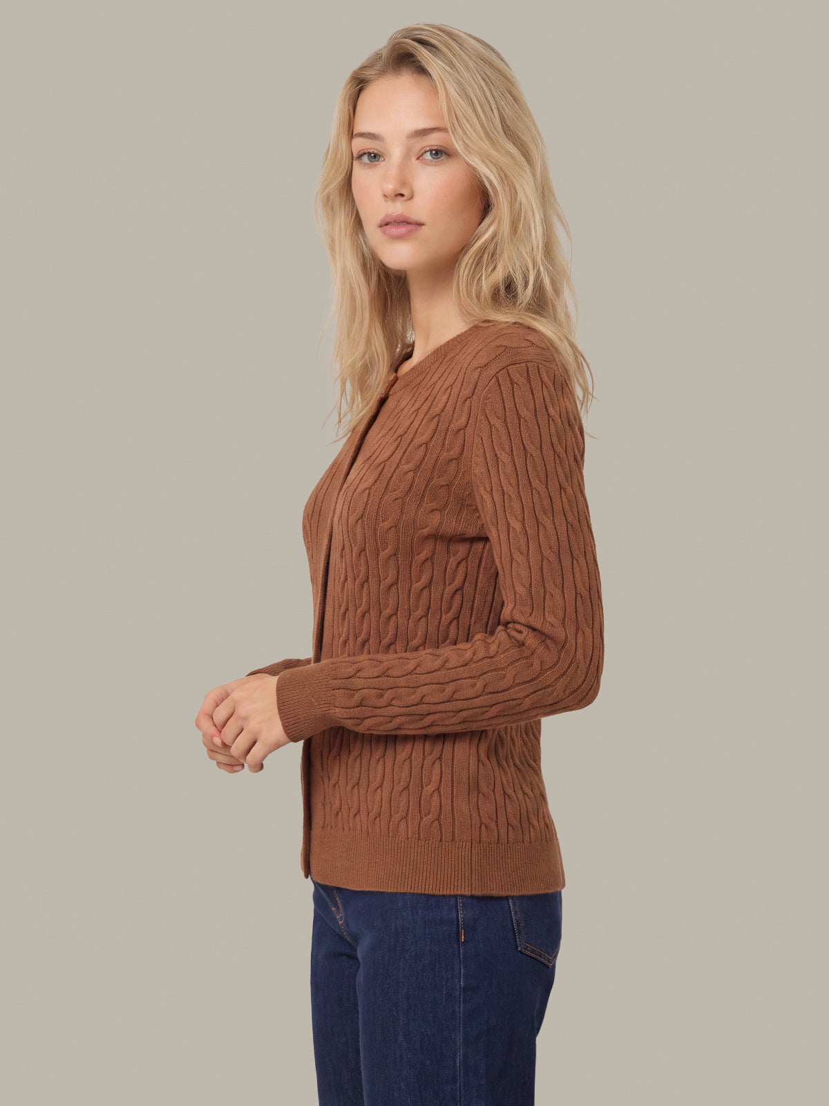 A woman wearing a brown cable-knit cardigan sweater and dark blue jeans, standing against a solid light gray background.