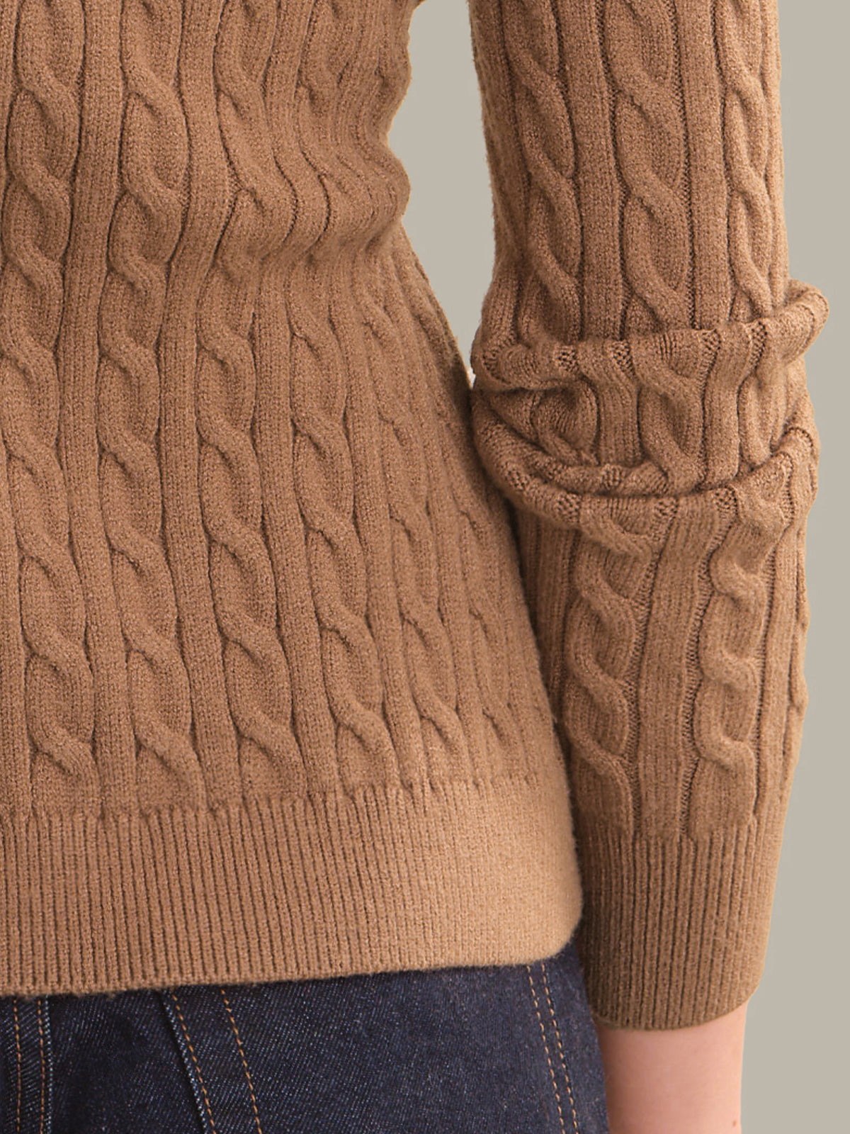 Close-up of a person wearing a camel brown cable-knit sweater with rolled cuffs, paired with dark blue jeans against a neutral gray background.