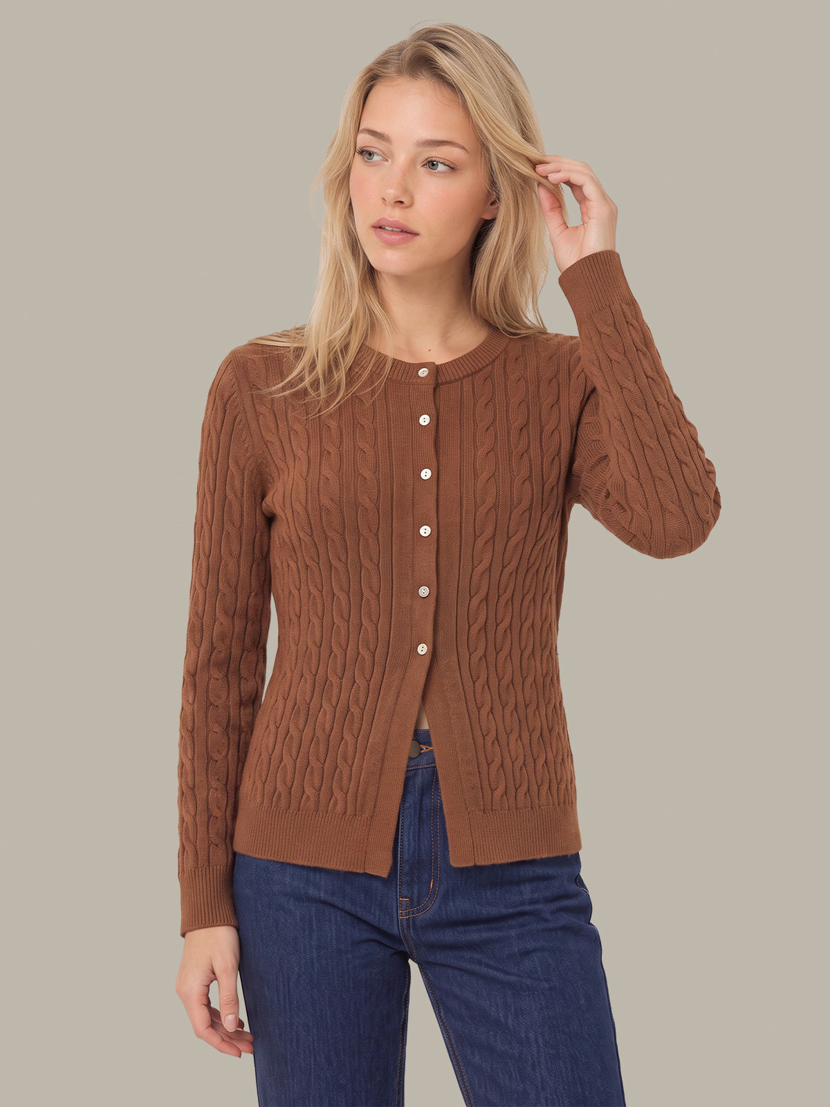 A woman modeling a brown cable-knit cardigan sweater with a button-up front, paired with blue jeans, against a solid gray studio background.