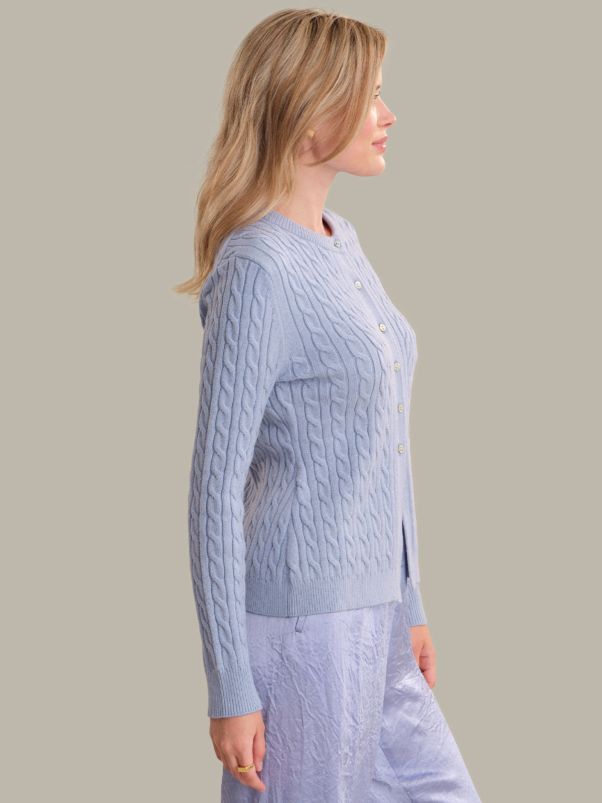 A woman shown in side profile wearing a light blue cable-knit cardigan with buttons and matching light blue textured pants, against a plain warm gray studio background.