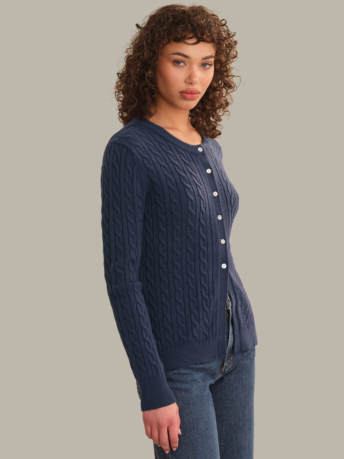 A woman with curly brown hair wearing a navy blue cable-knit cardigan sweater with white buttons, paired with blue jeans, against a solid light gray background.