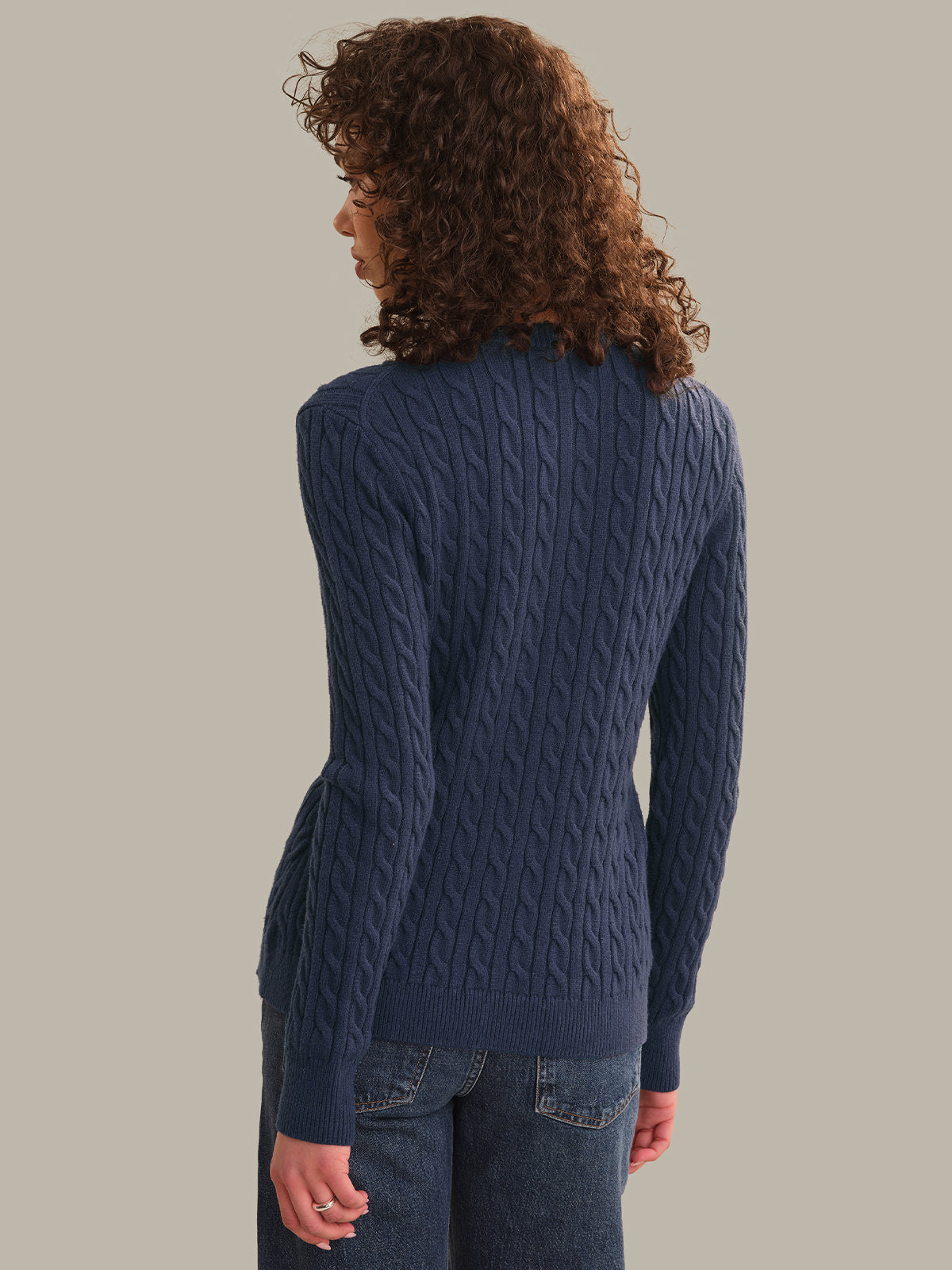 A woman with curly brown hair is shown from the back wearing a navy blue cable-knit cardigan and dark blue jeans against a solid light gray background.