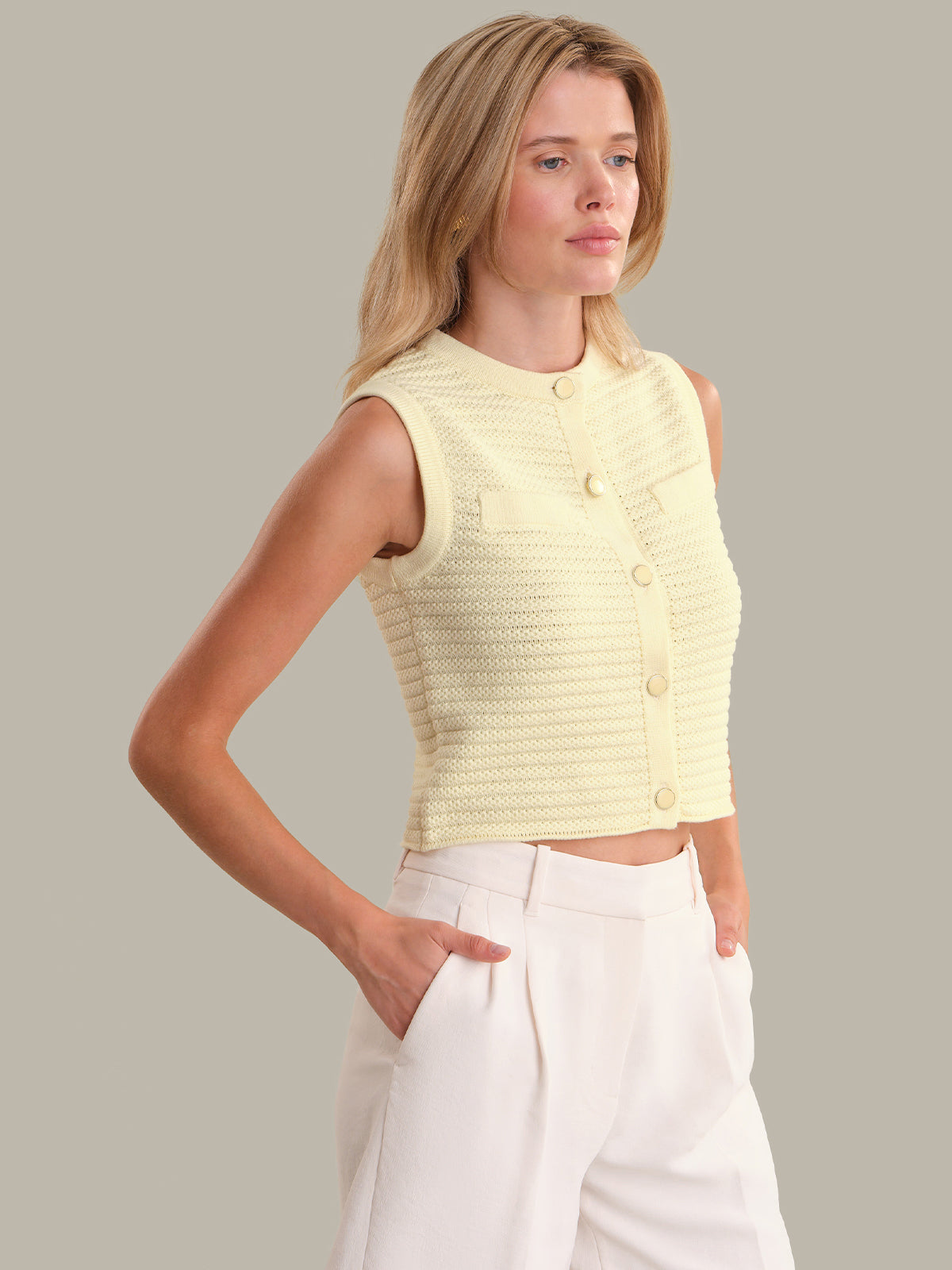 Woman wearing a light yellow sleeveless knitted top with buttons, standing against a plain background.