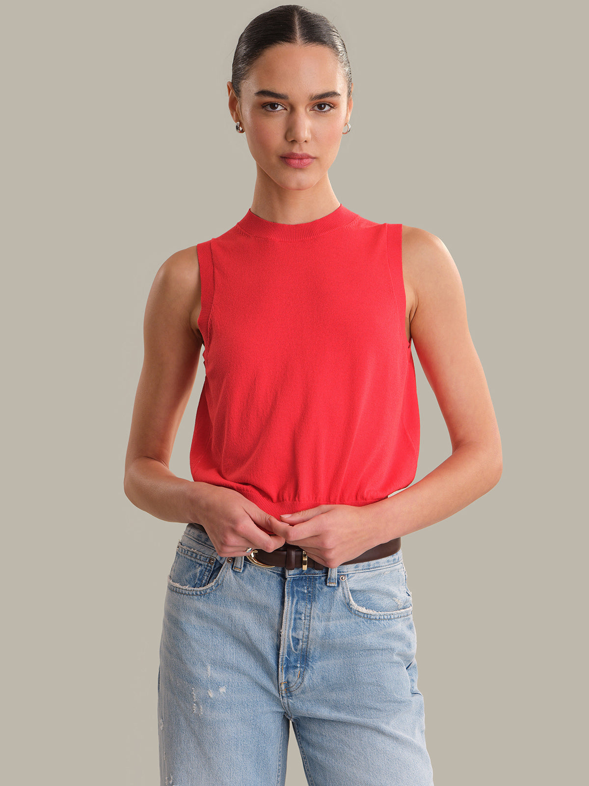 Woman wearing a red sleeveless top and light blue jeans against a gray background