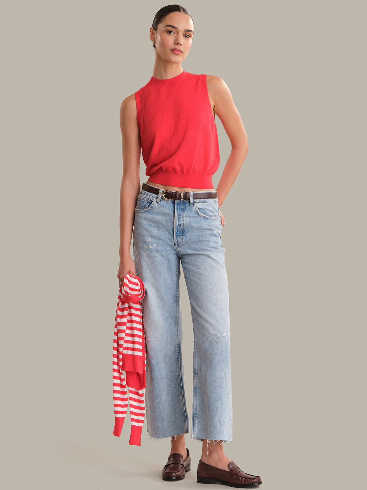 Woman wearing a red sleeveless top and light blue jeans, holding a striped shirt, on a gray background.