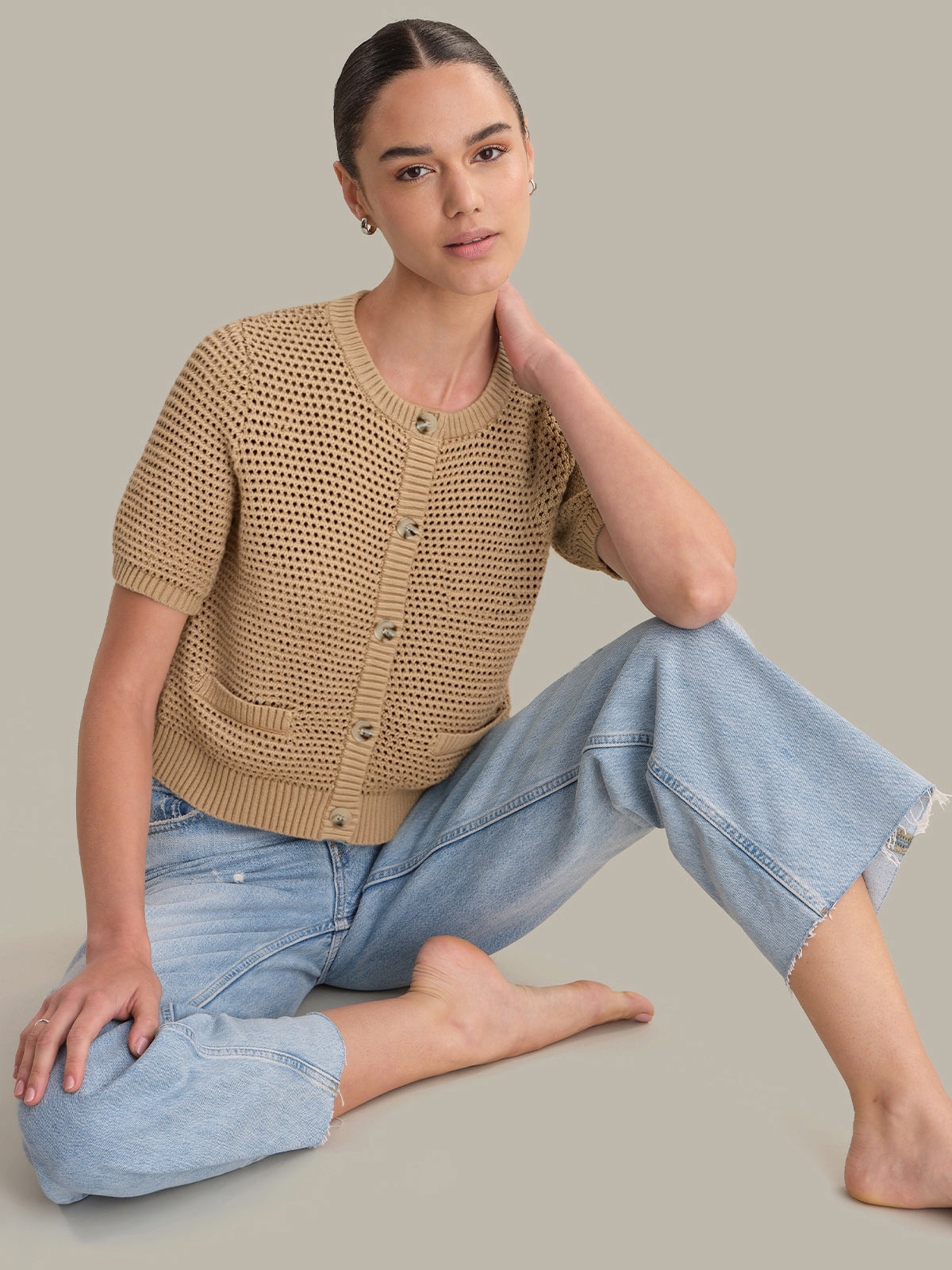 Woman wearing a beige knitted cardigan and light blue jeans sitting on a gray background