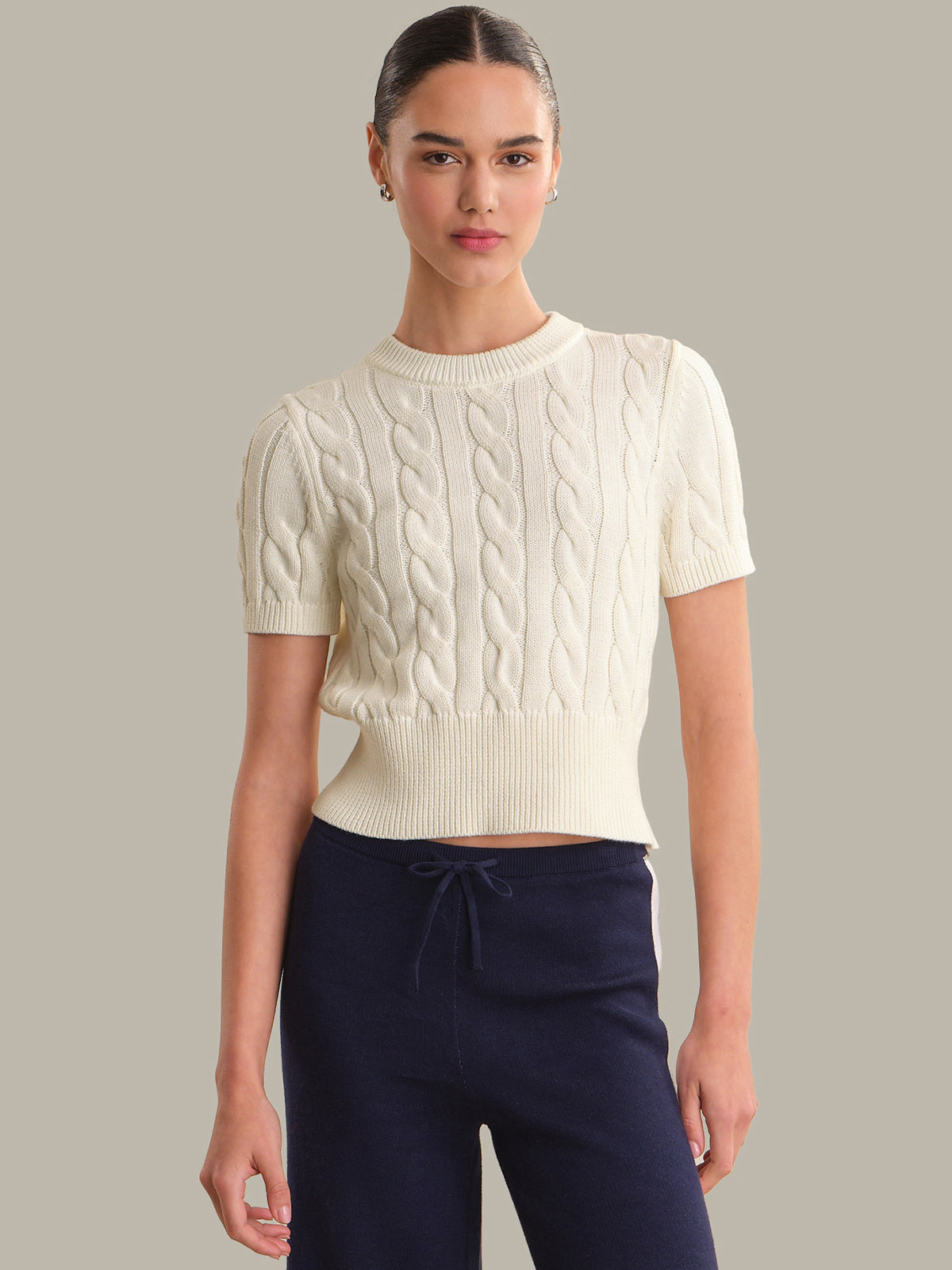 Woman wearing a cream cable knit sweater and navy pants against a neutral background