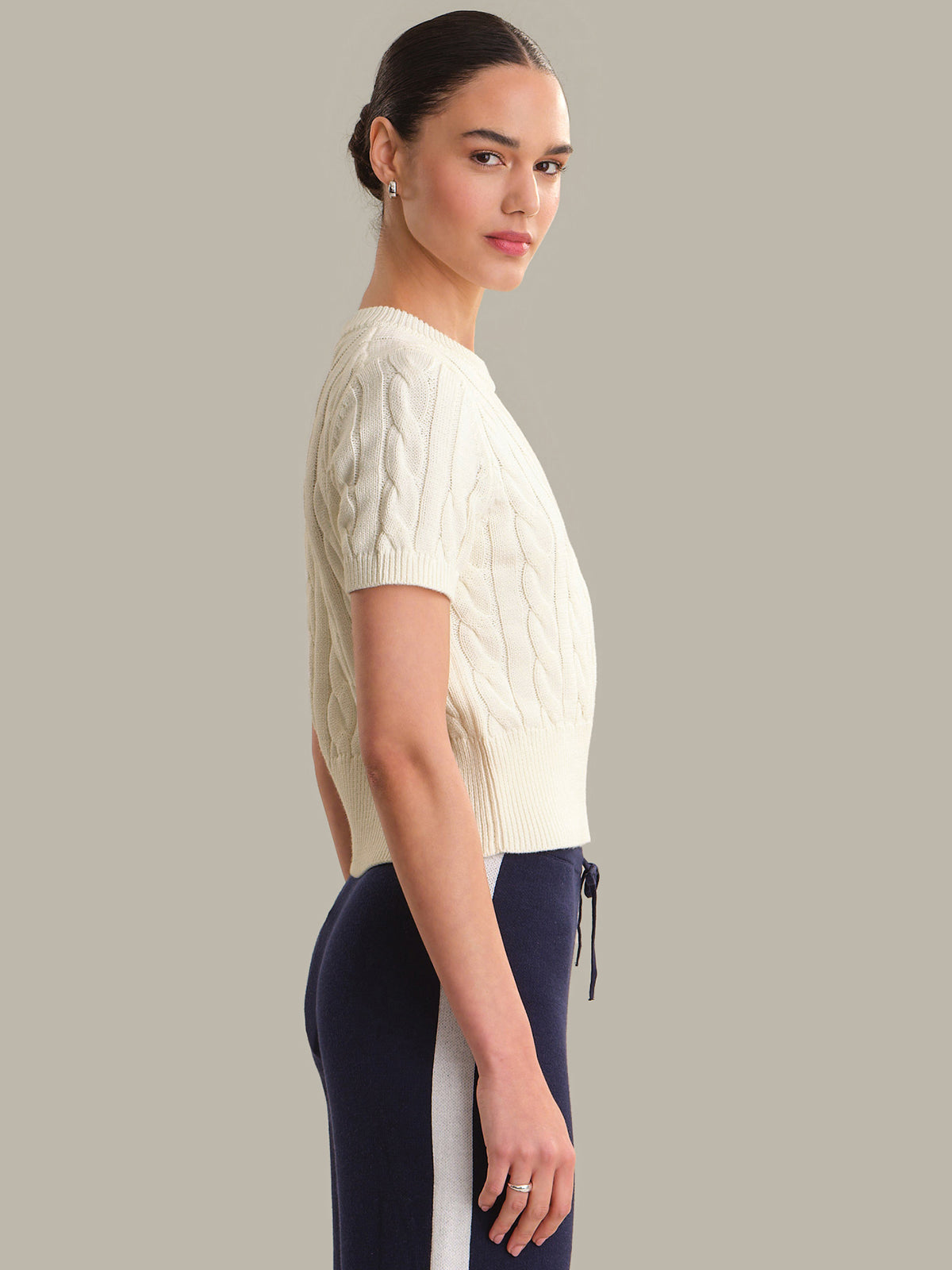 Woman wearing a cream cable knit sweater and dark pants on a gray background