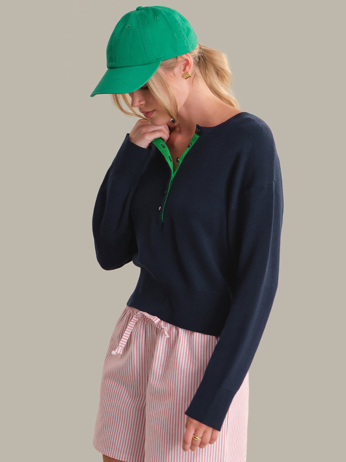Person wearing a navy sweater, pink shorts, and green cap on a gray background