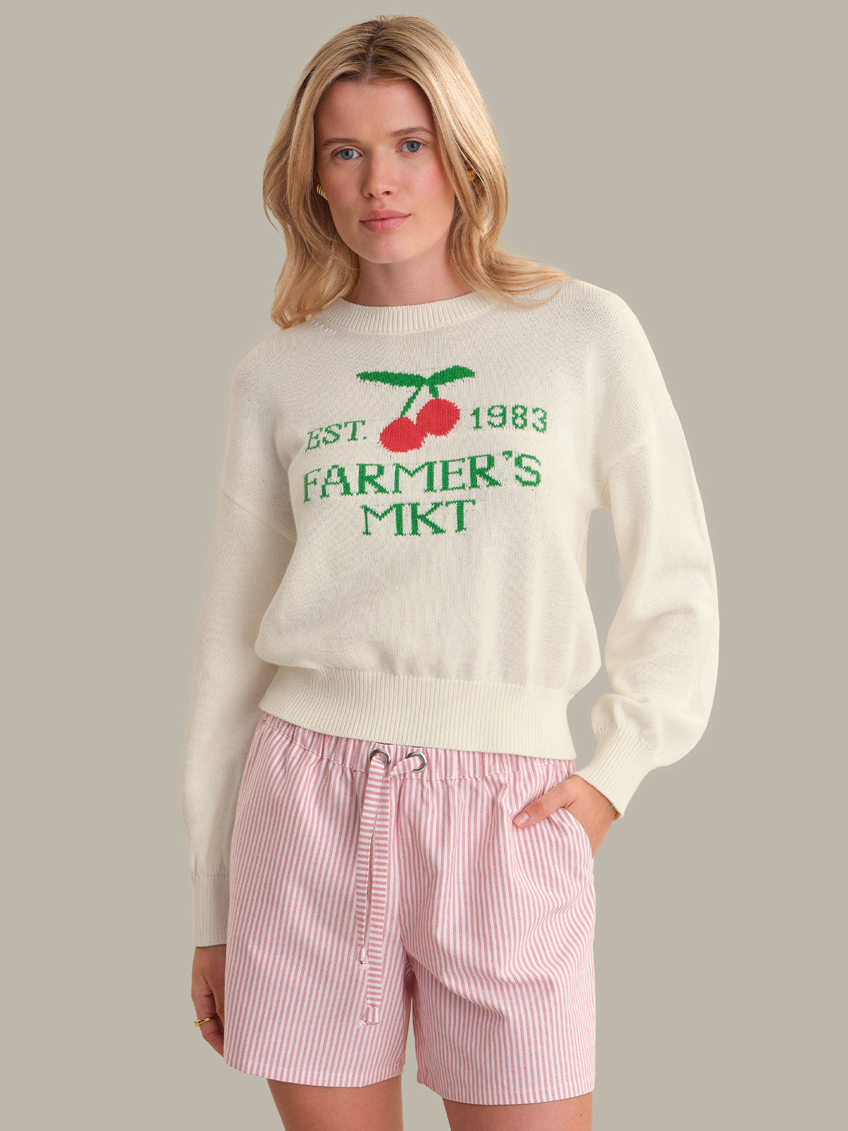 Woman wearing a cream sweater with text and cherries, paired with pink shorts on a gray background