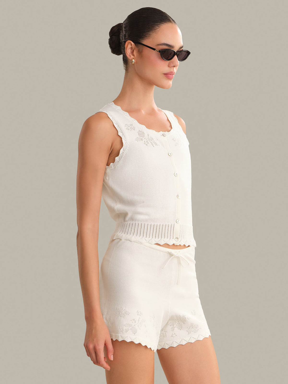 Woman wearing a white sleeveless top and shorts set with sunglasses on a gray background