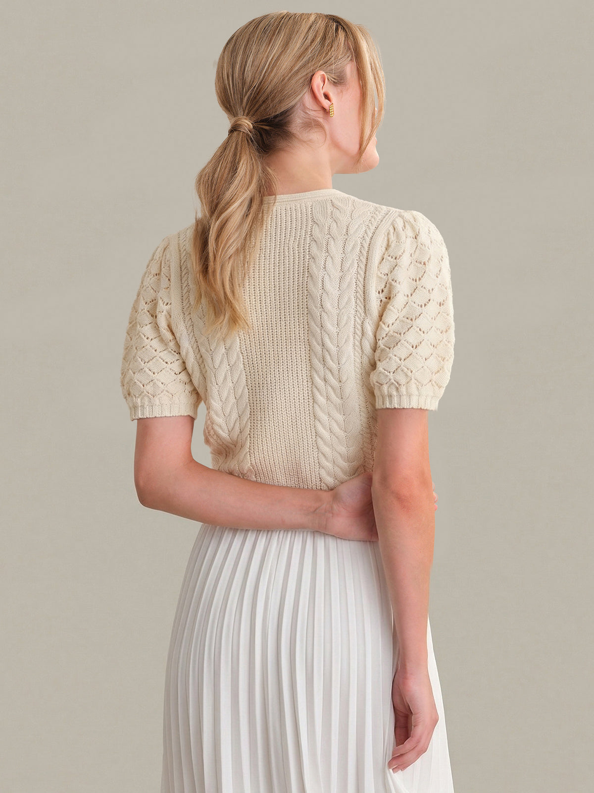Woman wearing a cream-colored knitted top and white pleated skirt against a plain background