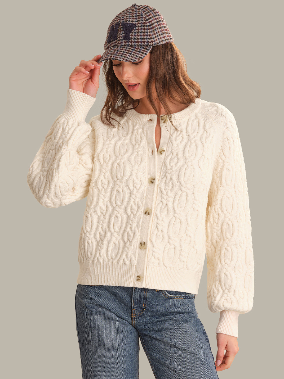 A woman wearing a cream cable-knit cardigan with gold buttons, blue jeans, and a plaid baseball cap, posed against a solid gray studio background.