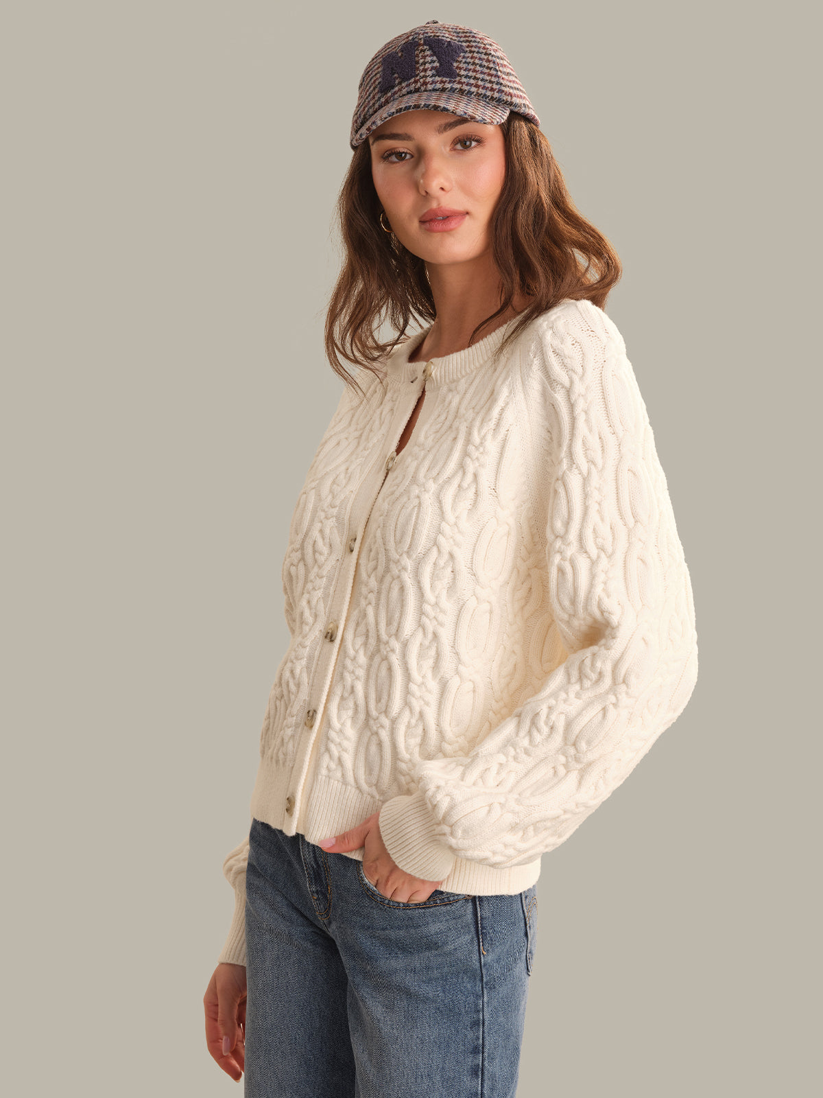 A woman wearing a cream cable-knit cardigan, blue jeans, and a plaid cap, photographed against a solid light gray studio background.
