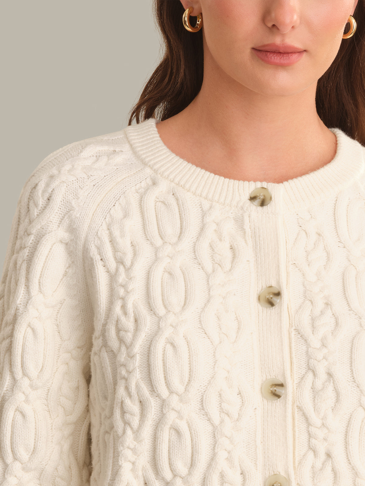 A close-up of a woman wearing a cream-colored cable-knit cardigan sweater with decorative buttons, set against a neutral light gray background.