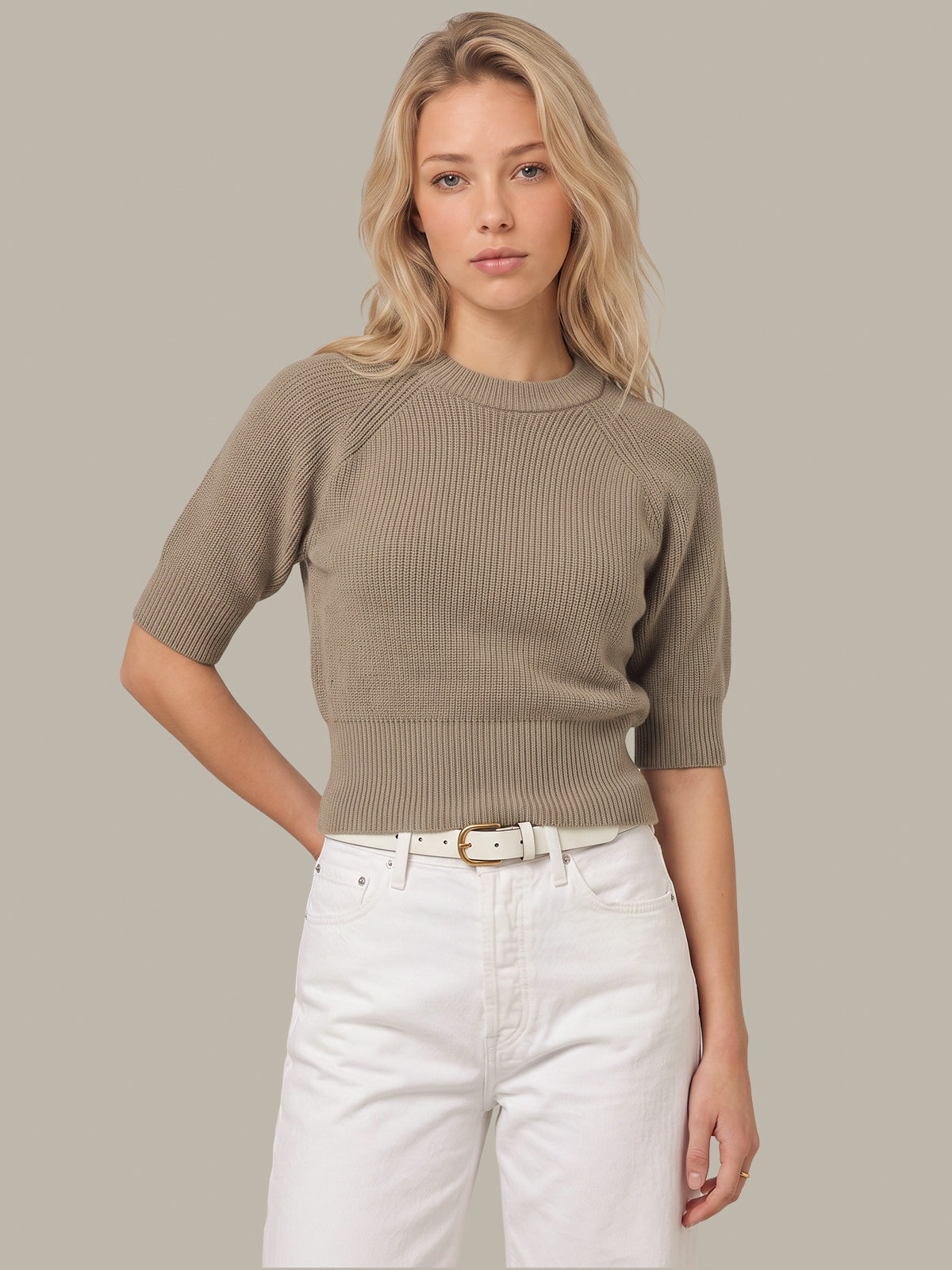 A blonde woman wearing a taupe ribbed short-sleeve crewneck sweater and white jeans, standing against a solid taupe background.