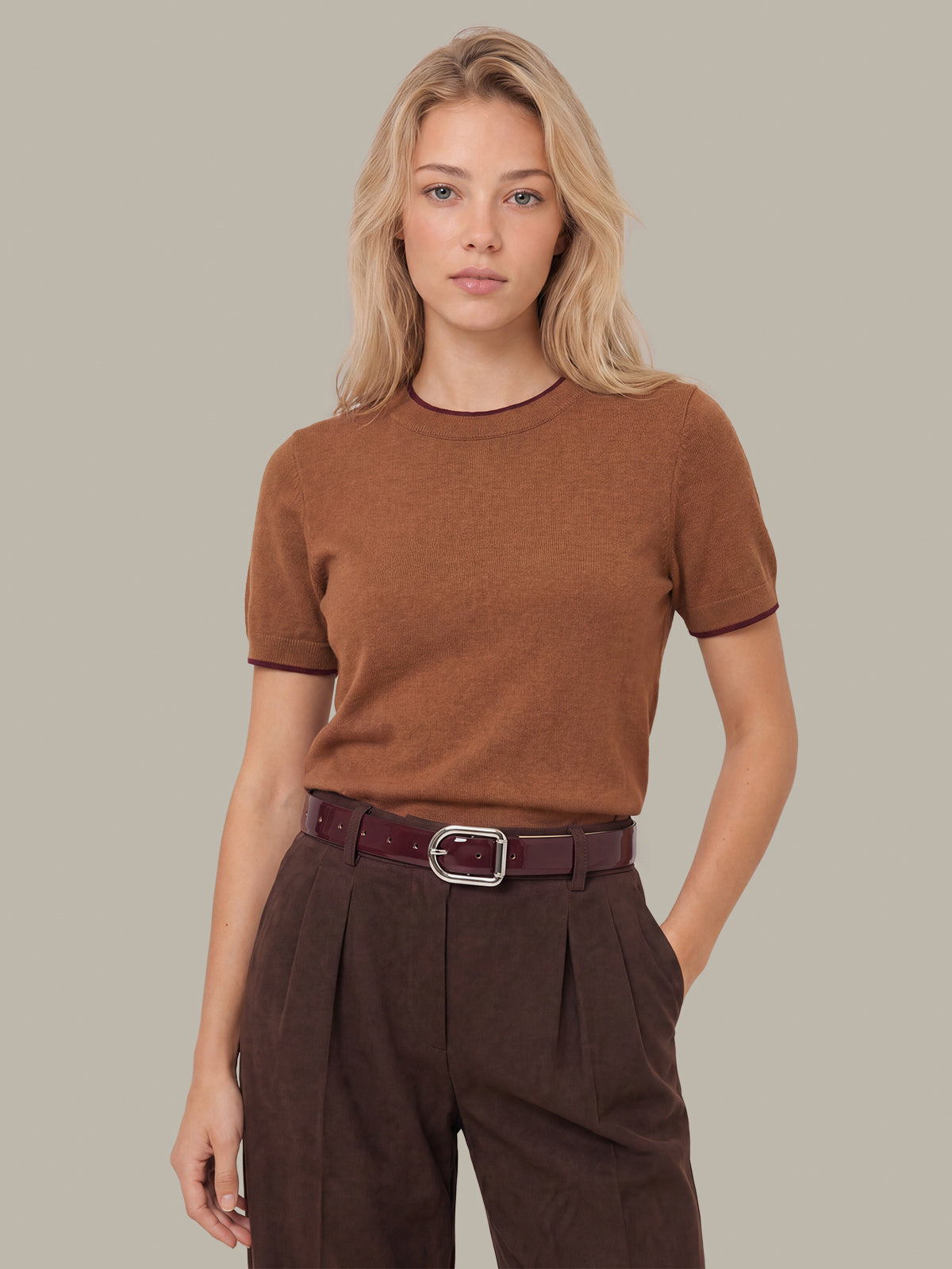 A woman modeling a short-sleeve crewneck knit top in a warm brown color, paired with dark brown pleated trousers and a dark red belt, against a solid light gray background.