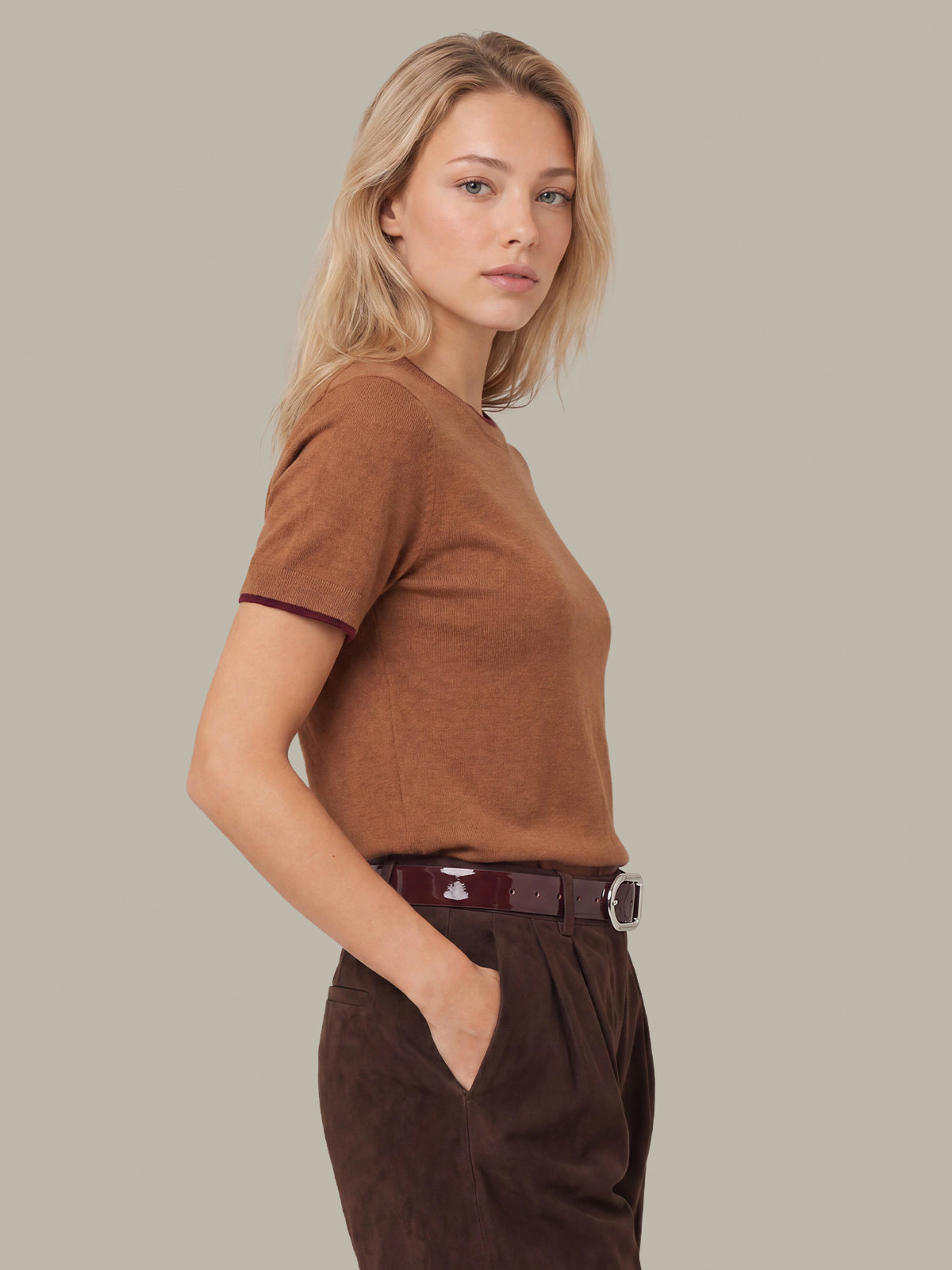 A woman wearing a fitted short-sleeve brown knit top and dark brown trousers, posed in profile against a plain warm gray studio background.